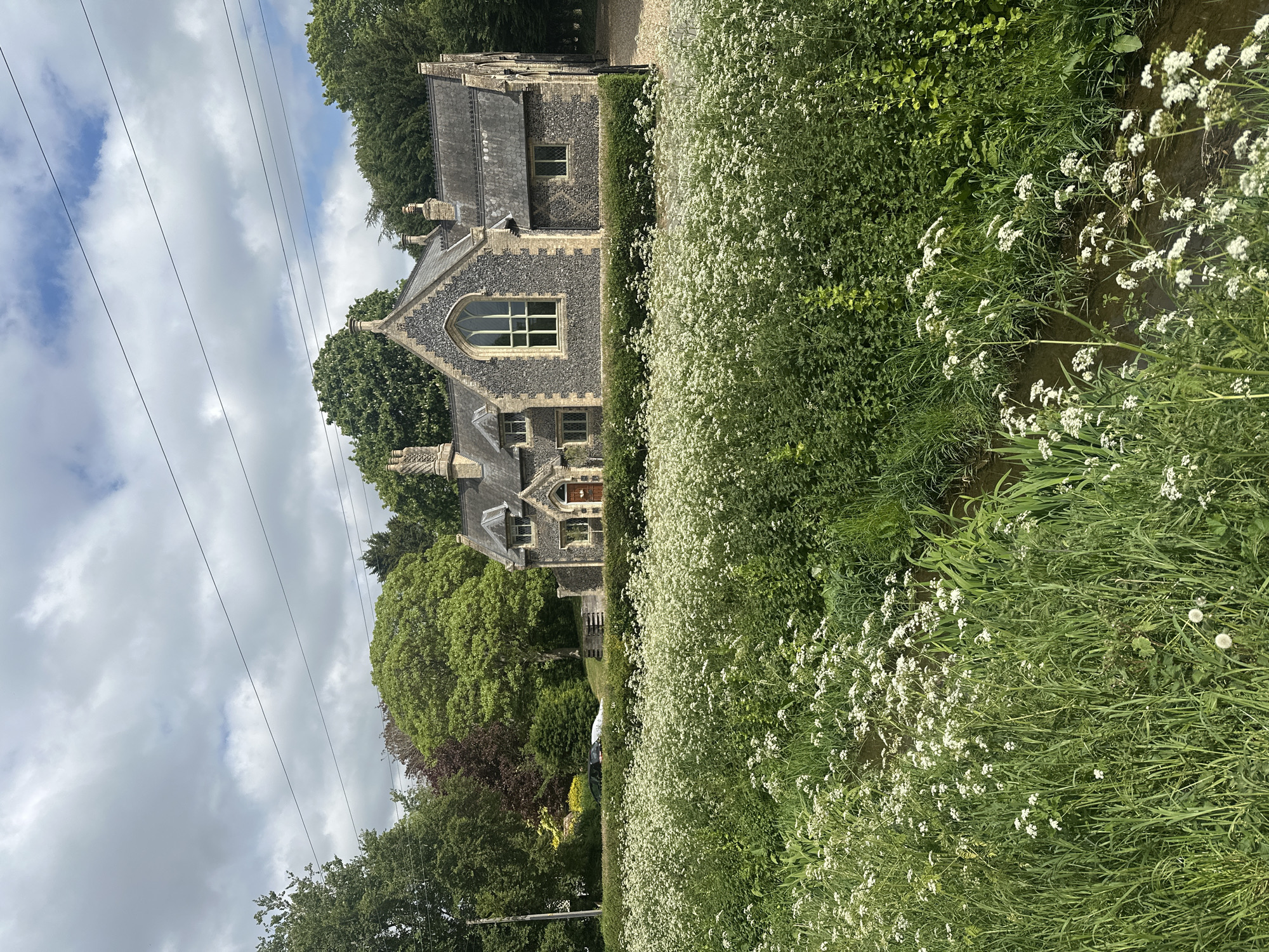 This is an outdoor shot of a building located in West Suffolk, United Kingdom. The building is gray stone, with a pointed roof and arched windows. A profusion of white flowers and green plants grow alongside the building and obscure part of the scene. Green trees surround the building. Overhead wires run above the trees. The sky is mostly cloudy.
