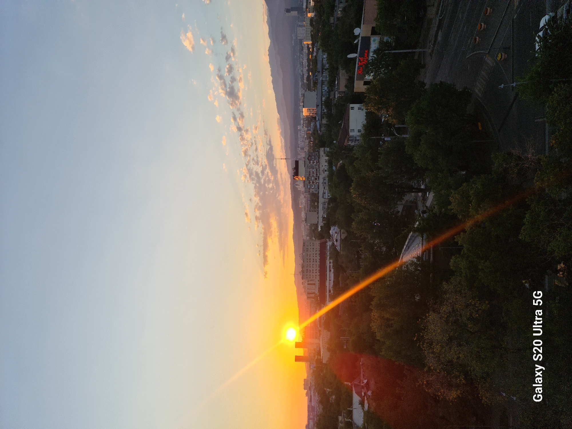 The image depicts a sunset over a city skyline in Almaty, Kazakhstan. The sun is low on the horizon, casting a warm orange glow and a prominent sunbeam that stretches diagonally across the frame. Buildings of varying heights, some appearing to be residential and others commercial, are scattered throughout the cityscape. Green trees form a dense canopy in the foreground and mid-ground, interspersed with roads and what appear to be streetlights. In the background, a range of mountains is visible, their shapes softened by the atmospheric haze of sunset. The sky transitions from a pale blue at the top to a vibrant orange near the horizon, with scattered, softly illuminated clouds. The words "Galaxy S20 Ultra 5G" are overlaid on the lower left corner of the image, indicating it was taken with a Samsung Galaxy S20 Ultra 5G smartphone. No people are visible in the scene.