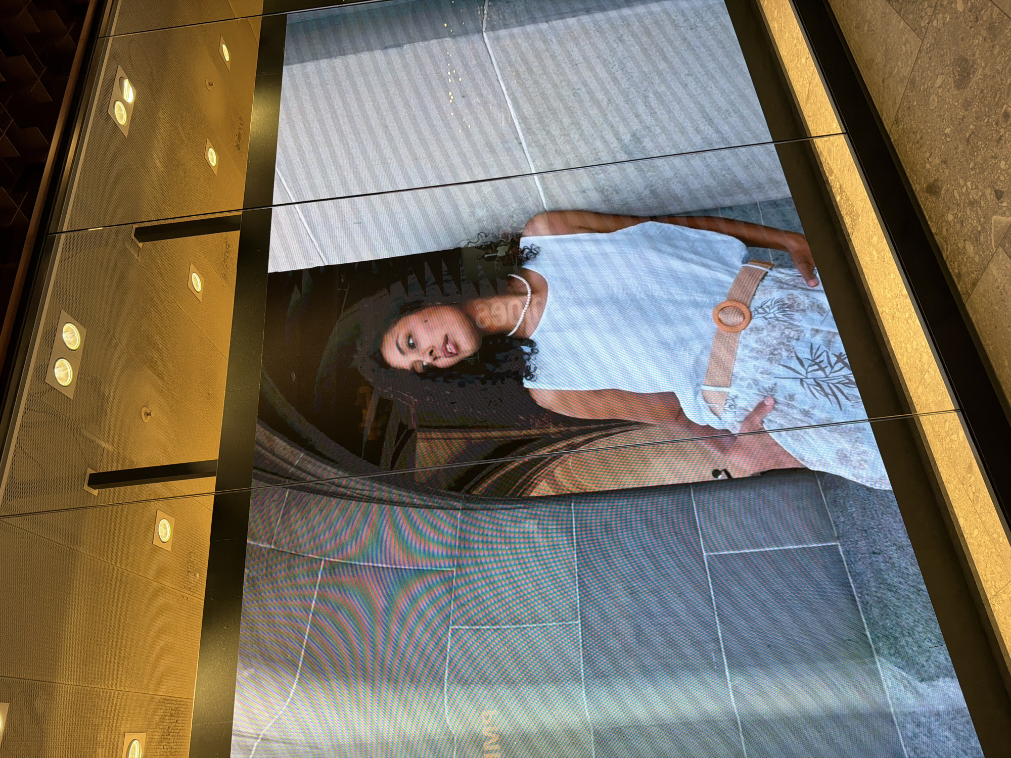 A digital display showcases a fashion advertisement featuring a young Black woman with curly hair standing against a stone wall. She is wearing a white tank top, a patterned skirt with a woven belt, and a pearl necklace. The display is framed by a dark border and positioned above a textured floor. The background of the advertisement depicts an arched stone structure, possibly a doorway or architectural feature. The lighting in the image appears to be artificial, with yellow-toned lights visible on the ceiling above the display. The overall scene suggests an indoor commercial setting.