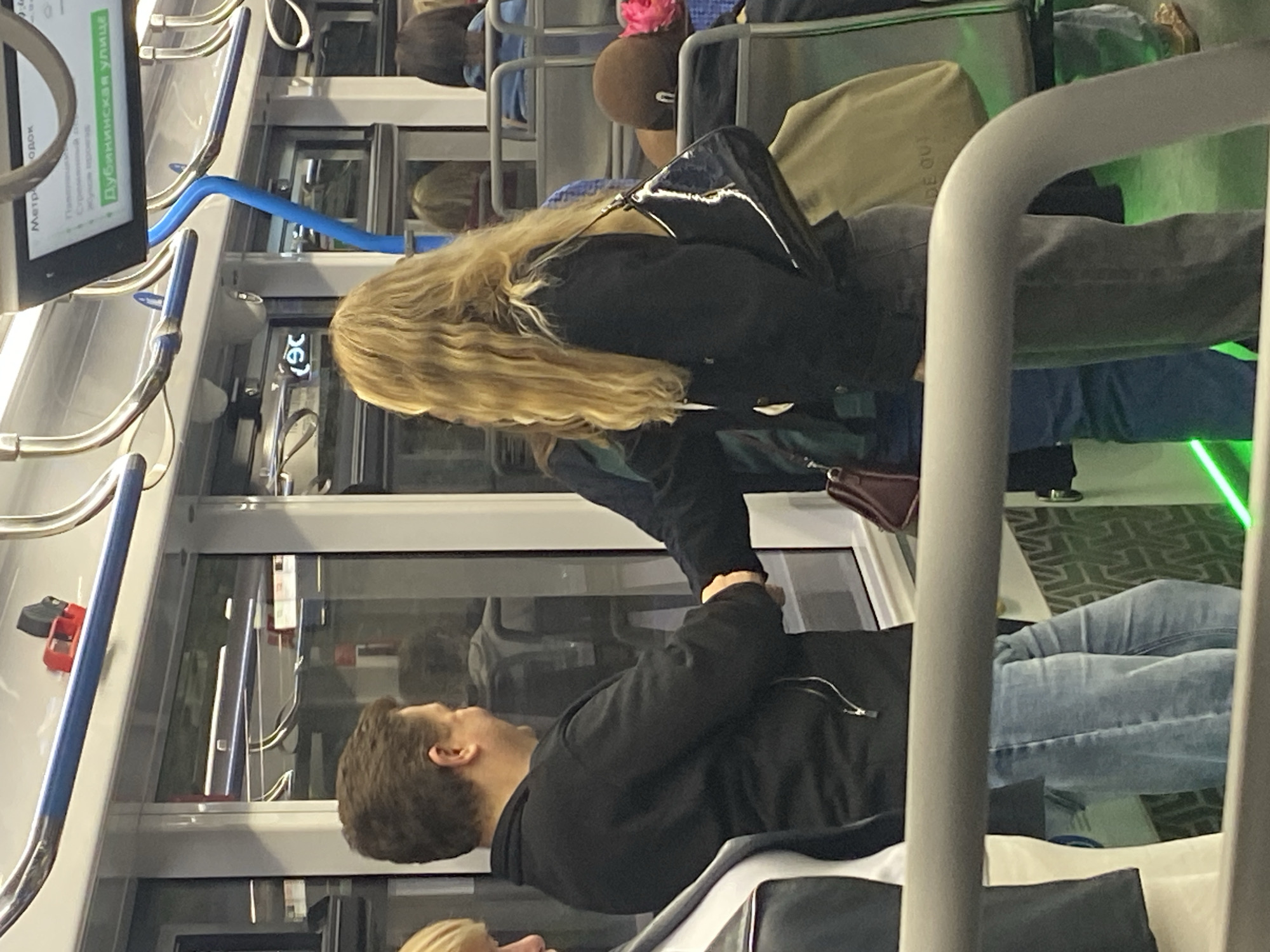 The image captures an interior scene of a public transportation vehicle, likely a metro or tram, in Moscow, Russia. Two individuals, a man and a woman, are positioned in the foreground. The man, wearing a black jacket and blue jeans, is facing the woman and appears to be engaged in conversation or an interaction. The woman, with long blonde hair, is wearing a black jacket and gray pants, and she is holding a black handbag with a shiny finish. Other passengers are visible in the background, seated or standing, and the environment includes handrails, windows, and digital displays with Russian text, one of which shows "Дубининская улица" (Dubininskaya street), indicating a stop or route. The overall lighting suggests an indoor, possibly evening or overcast, setting.