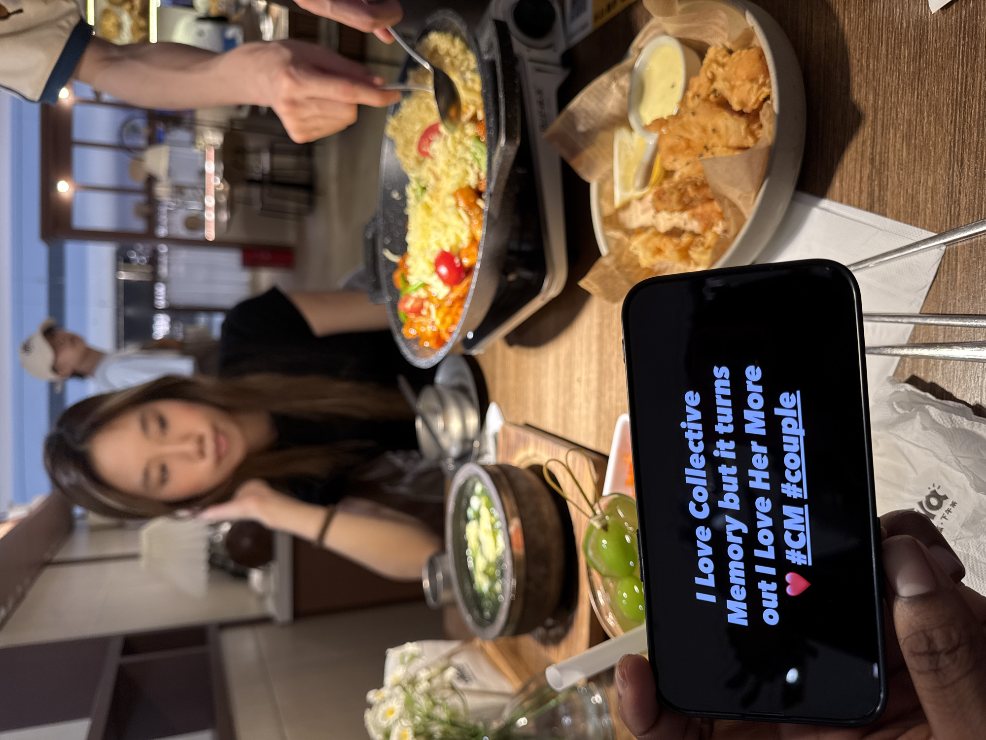 The image depicts a person holding a phone in front of a table filled with food. On the phone screen, the text "I Love Collective Memory but it turns out I Love Her More #CM #couple" is displayed. On the table, there is a cast iron pan with yellow noodles and vegetables, a bowl of fried food with a lemon wedge and dipping sauce, a small bowl of green vegetables, and a bowl of grapes. The person holding the phone has dark skin. A woman with long hair is sitting at the table looking down. Another person is standing and using a spoon to stir the food in the pan. The location appears to be a restaurant in Yuzhong District, China, as indicated by Chinese characters visible in the lower right corner of the image. There are other people and objects visible in the blurred background.