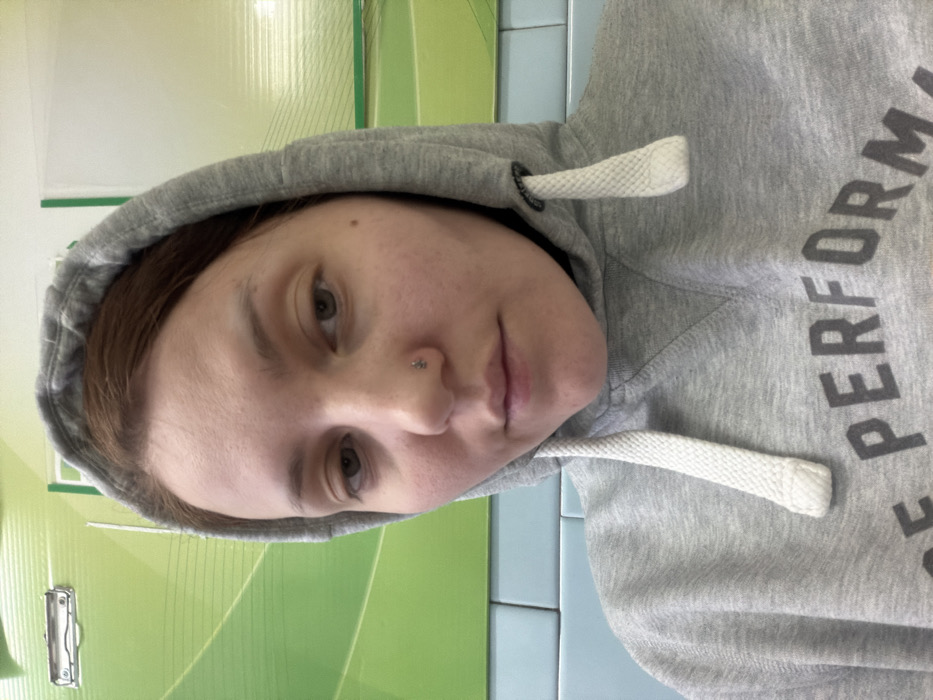 A person with light skin, brown hair, and hazel eyes, wearing a gray hooded sweatshirt, is photographed indoors. The hoodie's drawstrings are visible, and a silver nose piercing is on the left nostril. The background is a combination of green and blue surfaces. The person's face is slightly tilted, with a neutral expression. The image is rotated, presenting a sideways view.