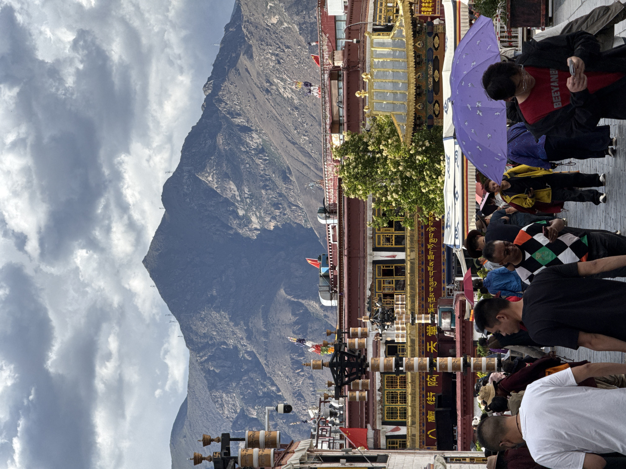 An outdoor scene captured under a cloudy, overcast sky showcases a bustling street or plaza with a majestic mountain range in the background. The image is dominated by traditional, ornate multi-story buildings with distinct Tibetan architectural elements, including golden roofs, red-painted walls, and decorative accents. In front of these structures, rows of cylindrical prayer wheels are visible, some featuring the swastika symbol (a Buddhist symbol of auspiciousness). Prayer flags and Chinese national flags flutter from the buildings. A security camera is also mounted near the buildings.
A crowd of people fills the foreground and midground. They are dressed in casual modern attire, engaged in various activities like walking and standing. One prominent figure, seen in a red t-shirt with the word "BEEYANE" printed on it, is looking at a mobile phone while partially shaded by a large purple umbrella adorned with bird patterns. Other individuals in the crowd include a person wearing an argyle-patterned top and another in a white t-shirt seen from behind. A tree with white blossoms adds a touch of nature amidst the structures. The massive, rugged mountain in the distance forms a dramatic backdrop, with faint structures, possibly transmission towers, visible on its slopes. The overall atmosphere suggests a lively day in a culturally rich area within the Chengguan District, China.