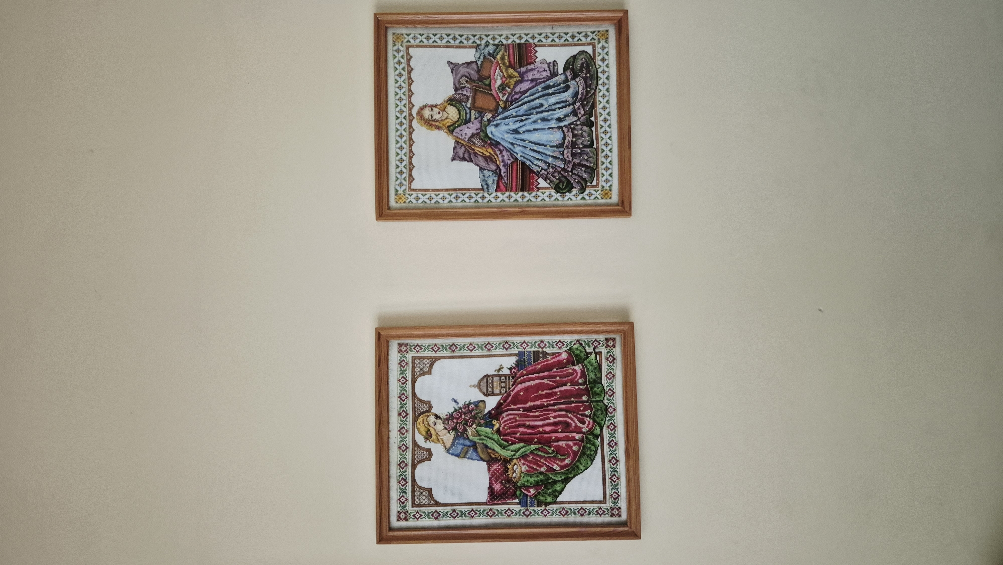 Two framed cross-stitch artworks of medieval women are displayed on a plain, light-colored wall. The artwork on the left depicts a woman in a red and green gown holding a bouquet of pink flowers, with a small birdcage visible to her right. The artwork on the right shows a woman in a blue and purple gown reading a book, with several cushions behind her. Both frames are made of light-colored wood. The scene is indoors.