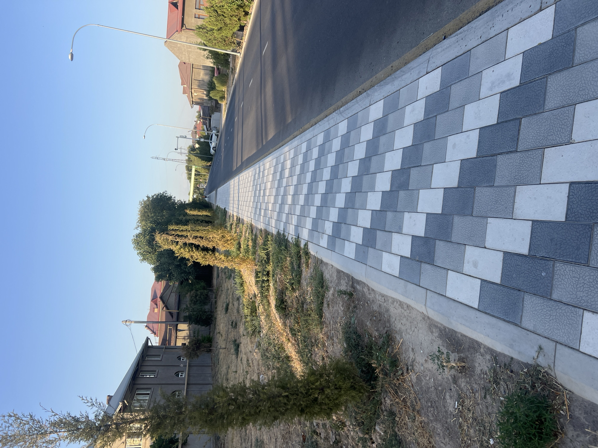 The image shows a suburban street scene on a clear day. A paved sidewalk with a patterned design of gray and white tiles runs along the left side of the asphalt road. Several slender trees and bushes line the sidewalk and the adjacent undeveloped land, which is covered in dry grass and dirt. On the far side of the road, houses with red-tiled roofs and gray walls are visible, interspersed with trees and vegetation. Streetlights are positioned along the road. A white car is parked on the right side of the road. The sky is a clear, pale blue.