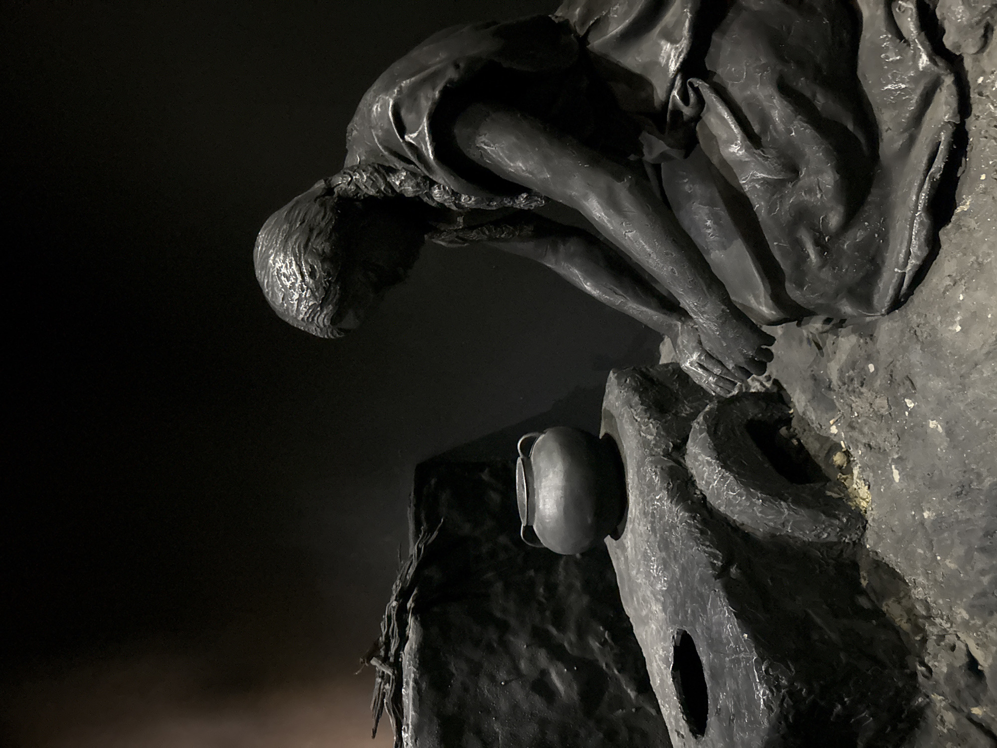 The image depicts a monochromatic diorama of a historical scene, likely within a museum exhibit. In the foreground, a sculpted figure of a person, appearing to be a woman with braided hair, is kneeling and focused on a hearth. The hearth is constructed of stone and has an opening at the front, with debris on the ground beneath it. A dark-colored pot with two handles sits atop the hearth structure. To the left, there is a pile of what appears to be firewood or branches. The background is dark and nondescript, with a subtle smoky or dusty haze on the far left. The overall impression is one of a domestic or utilitarian scene from a past era. This diorama is located in the Tamar Regional Council, Israel, specifically at the Karkur museum, which is part of the "Museums of the Tamar Regional Council" complex.