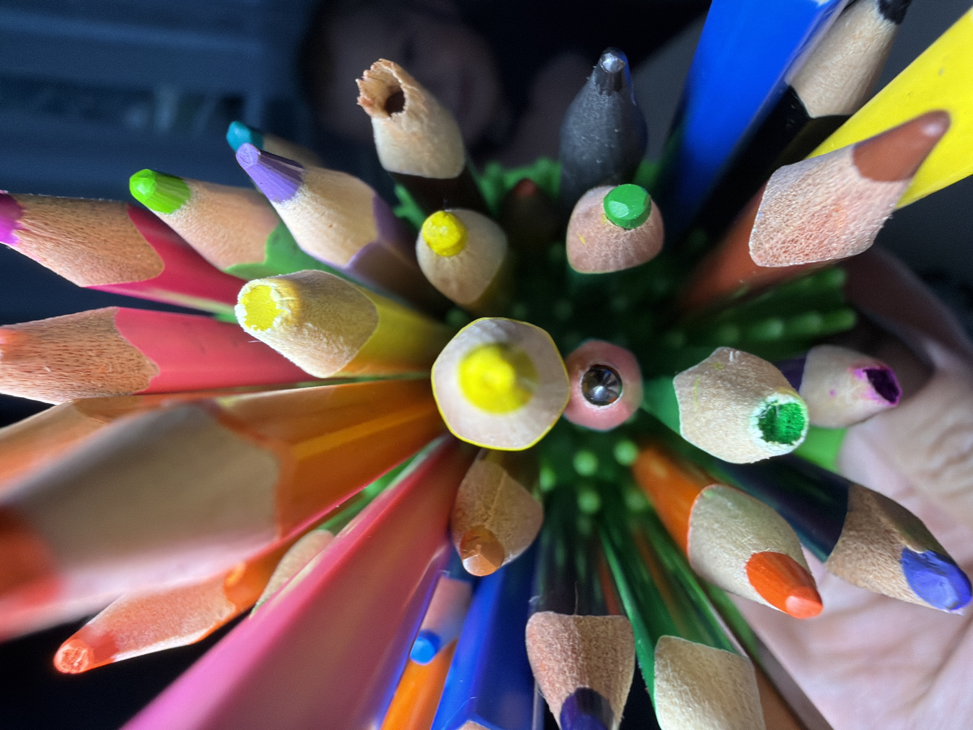 The image is a close-up, macro photograph of a cluster of sharpened colored pencils, fanned out and pointing upwards. The pencils are in various colors including pink, orange, yellow, green, purple, blue, black, and brown. Some of the pencil tips show the wood grain and the colored lead. The background is dark and out of focus, with a hint of a person's face visible in the upper right portion of the image. The lighting is dramatic, creating highlights and shadows on the pencils. The scene appears to be indoors, possibly a home or a classroom, with no specific landmarks of Votkinsk, Russia, visible.
