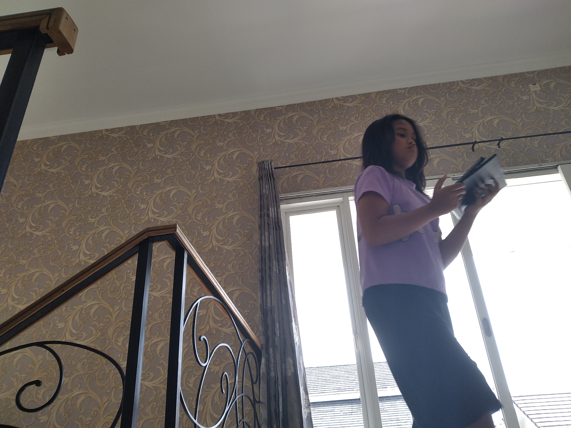 A low-angle shot captures a young girl in a purple t-shirt and dark skirt standing indoors, holding a tablet device. She is positioned to the right of the frame, with her body angled towards the left. Behind her, a large window with a white frame is visible, revealing a bright, overcast sky and the roof of a building outside. Dark, floral-patterned curtains hang beside the window.
The foreground features a section of a wooden and metal railing with ornate scrollwork, suggesting an indoor staircase or balcony. The railing is made of black metal with a light brown wooden top rail. The wall behind the railing is adorned with textured, ornate wallpaper in shades of beige and gold, featuring a repeating damask pattern.
The scene is lit by natural light coming from the window. The overall impression is of an indoor domestic setting, possibly a home, in Kaliwates, Indonesia. There are no other individuals or animals visible in the image. The girl appears to be focused on the tablet she is holding.
