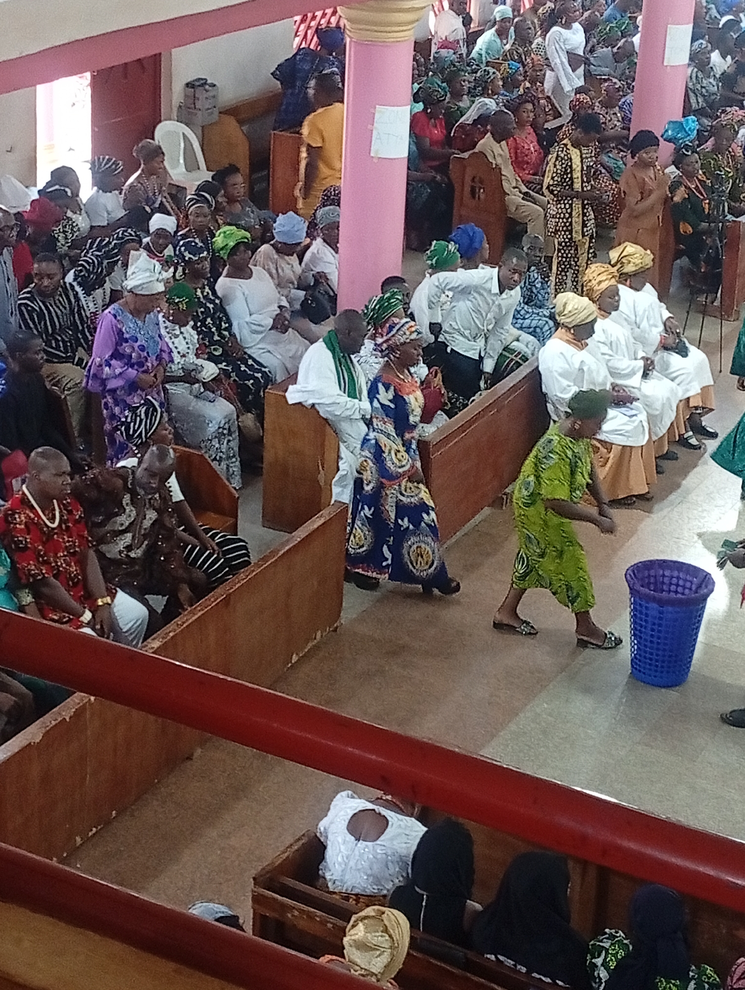 Captured at an undisclosed location within the city of Unknown, Unknown, the image presents a gathering of numerous individuals inside a church-like setting. The people, largely adorned in traditional African attire, populate pews and seating arrangements. The environment features pink-colored pillars, wooden pews, and a red banister at the forefront. Actions include walking and sitting; interactions are communal. Objects include a blue plastic basket and assorted personal items carried by the attendees.