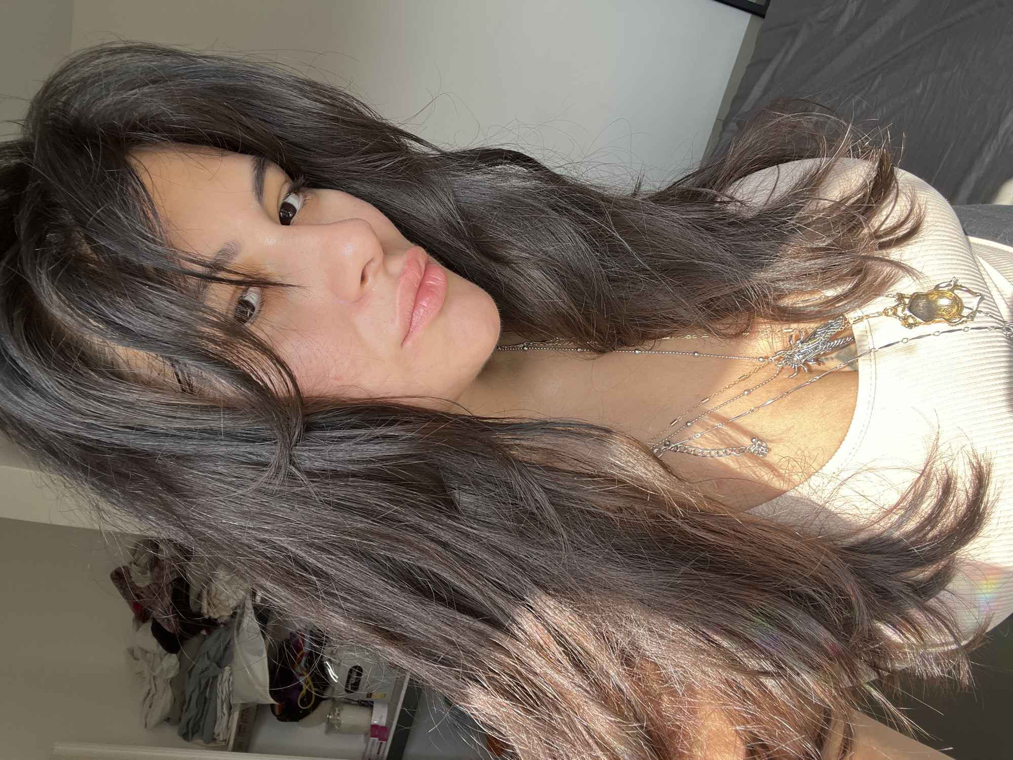 A woman with long, dark, wavy hair, styled with bangs, poses for a selfie. She is wearing a light-colored ribbed tank top and multiple layered necklaces, including one with a beetle pendant and another with a charm. The lighting suggests it is daytime, with sunlight casting a warm glow and creating some lens flare on her hair. In the background, on a shelf, there are various items including what appear to be candles and folded laundry. The setting appears to be an indoor residential environment. There is no discernible indication of a specific location in Kiryat Bialik, Israel.