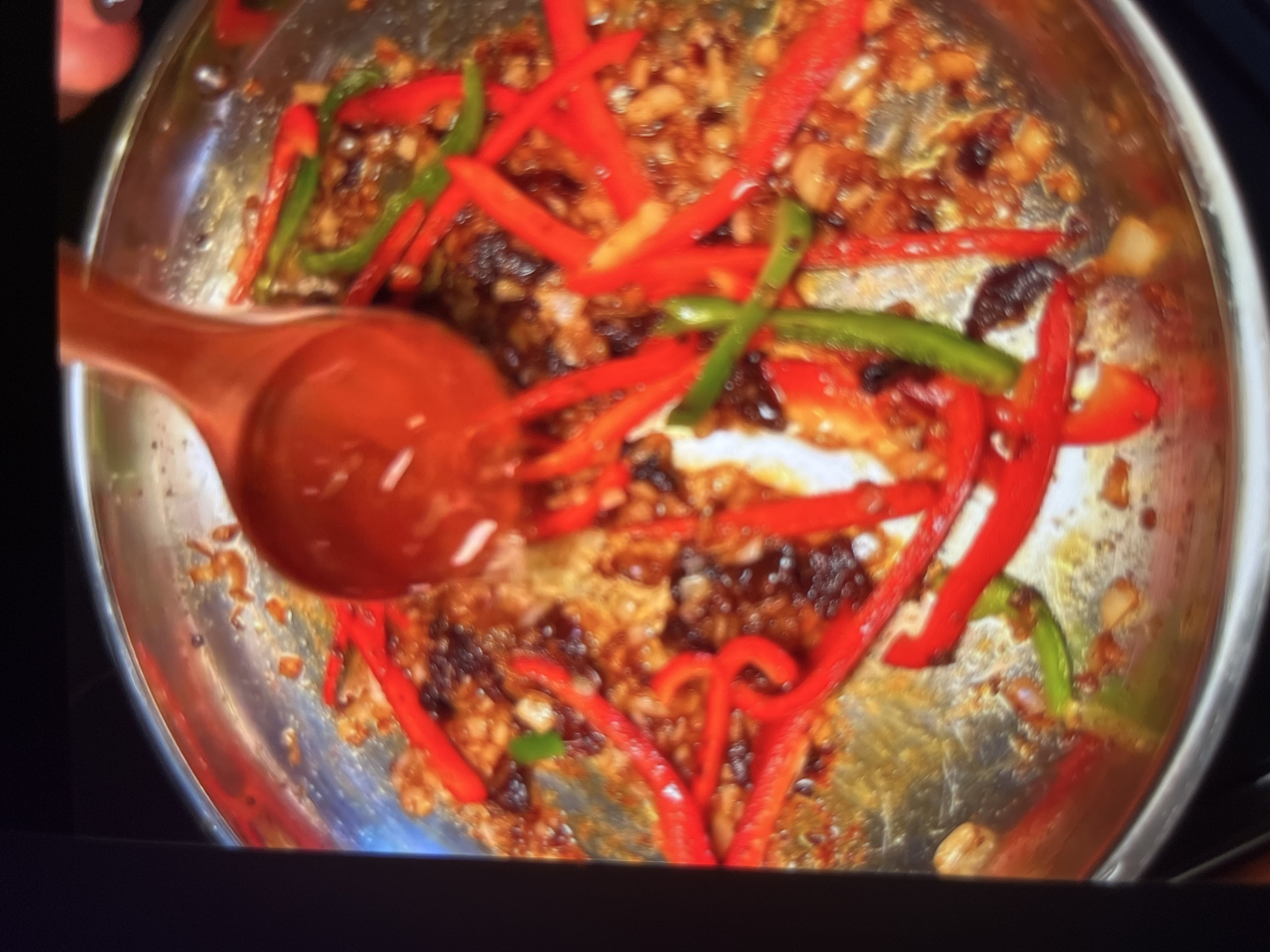 A close-up, blurry, overhead shot captures a stainless steel pan filled with food. The pan is on a stovetop, though the stovetop itself is not visible. Red and green bell pepper strips are scattered throughout the pan, mixed with a dark, glossy sauce and what appear to be small pieces of garlic or onion. A large, wooden ladle with a red handle is partially submerged in the sauce, scooping up some of the ingredients. The lighting is warm and creates reflections on the pan's surface. There are no people visible in the image. The scene is a kitchen environment, likely in Abuja, Nigeria, given the context of the prompt.
