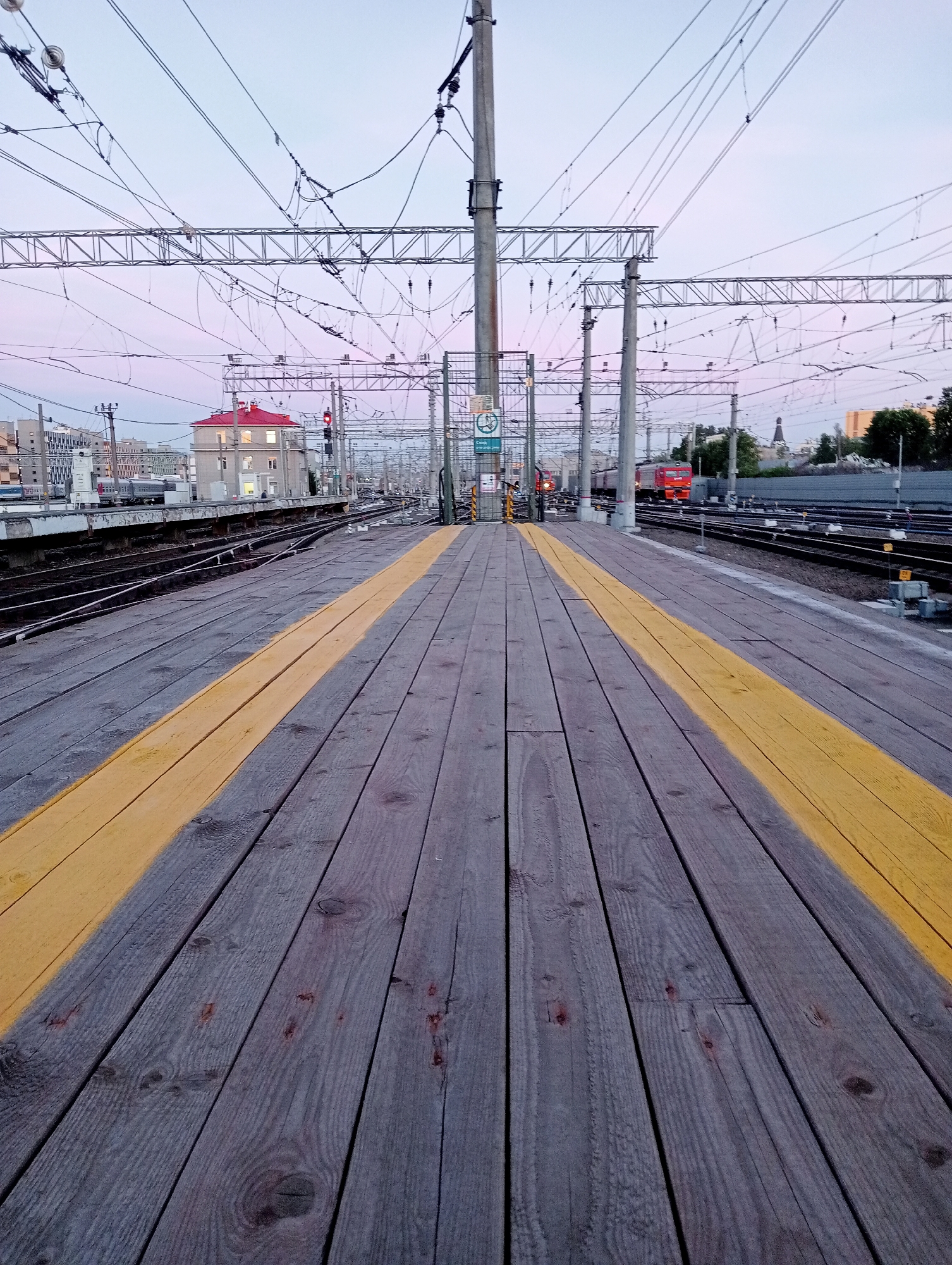 The image depicts a railway platform at dusk. The perspective is from the wooden platform itself, with two thick yellow lines marking a safety zone. In the background, multiple sets of railway tracks run parallel, crisscrossed by overhead electrical wires and supported by tall poles. A red train is visible on one of the tracks further down the line. On the left side of the frame, a building with a red roof and lit windows stands out against the pale sky. The overall scene suggests a functional, active railway station environment. The building with the red roof and the architectural style of the other buildings in the background are characteristic of Saint Petersburg, Russia. The presence of overhead electrical wires is typical for electrified railway lines common in Russia.
