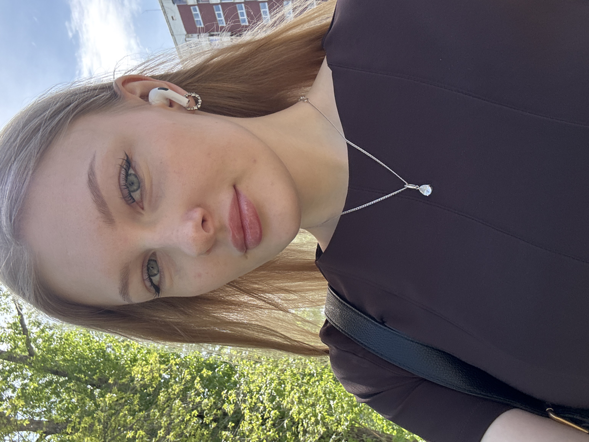 A young woman is posing for a selfie outdoors. She has fair skin, green eyes, and light brown hair. She is wearing a dark brown dress and a silver necklace with a teardrop pendant. She has a white wireless earbud in her left ear and a silver earring on the same ear. A black leather strap of a bag crosses her body. The background shows trees and a building, indicating an urban setting. Although I am unable to confirm location details, elements of the image are consistent with a city environment.