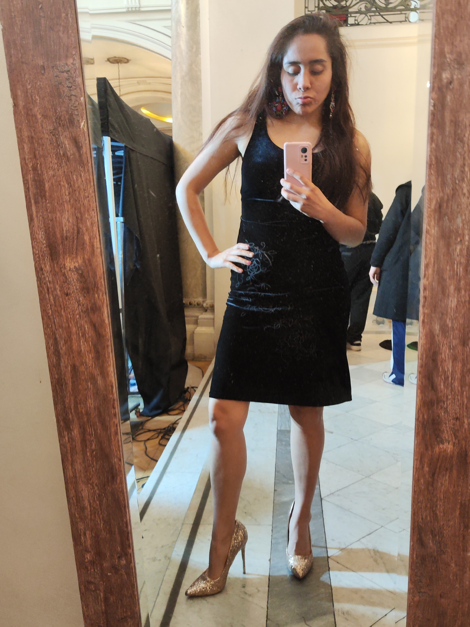 In a dimly lit indoor space, likely a museum or gallery with marble floors and classical architectural elements, a young woman with long, dark hair stands in front of a large, wooden-framed mirror. She is wearing a sleeveless, black velvet dress adorned with a subtle floral pattern and bright, colorful earrings. Her right hand is on her hip, and she holds a pink smartphone in her left hand, taking a selfie. She is wearing glittery, gold stiletto heels. In the reflection of the mirror, other people and a dark tarp covering equipment are visible, suggesting a behind-the-scenes or event setup. The woman's pursed lips and tilted head indicate a playful or slightly dramatic pose.