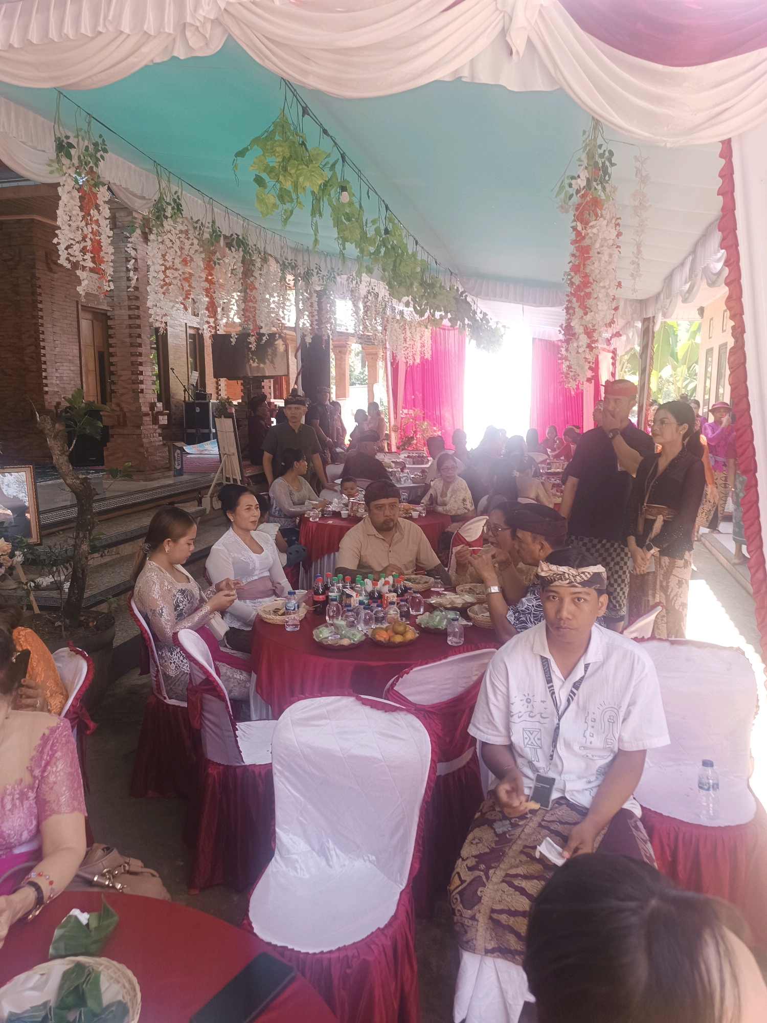 The image depicts an outdoor event, possibly a wedding reception, taking place under a decorated canopy in Penebel, Indonesia. Several tables are set up with red tablecloths and chairs draped in white and red fabric. Guests, dressed in traditional attire, are seated at the tables, some of which are laden with food and drinks. Floral arrangements, including hanging white and orange blossoms, adorn the canopy. In the background, a brick building with stairs and a doorway is visible. The lighting suggests it is daytime, with sunlight filtering through the canopy. The overall atmosphere appears festive and communal.