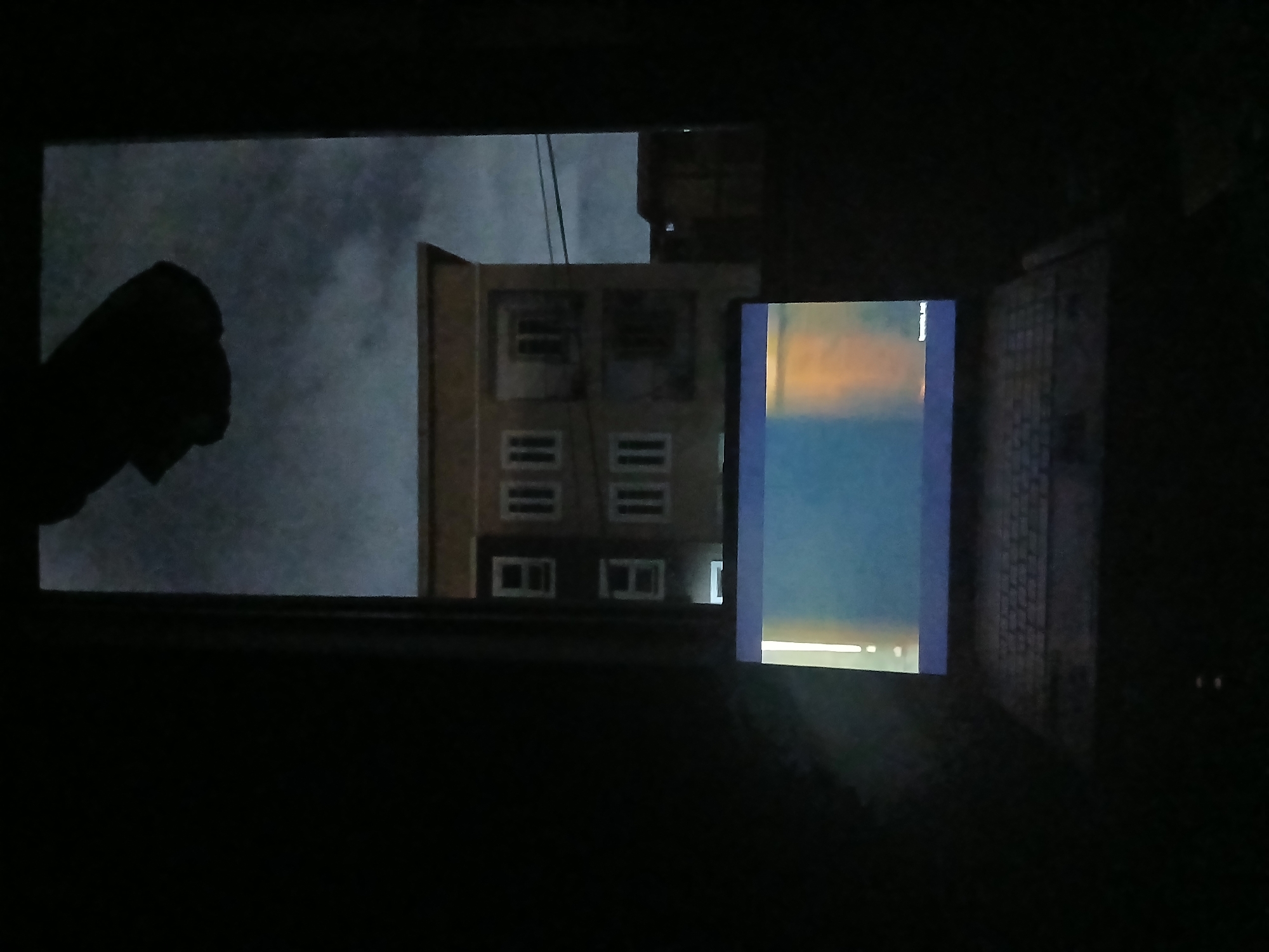 The image depicts an interior scene looking outwards through a window at night, focusing on a laptop in the foreground. The setting is consistent with an urban residential or mixed-use area in Lagos, Nigeria.
In the immediate foreground, a laptop with a visible keyboard is positioned, its screen illuminated. The screen displays a blue-to-orange gradient with a prominent vertical light-colored line on the left side, and faint, unreadable text in the bottom right corner. The surrounding interior is dark, indicating minimal or no internal lighting beyond the laptop screen.
Through the window, an exterior nighttime scene is visible. A multi-story, light-colored building with multiple rectangular windows is prominently situated directly opposite the vantage point. This building has a flat roofline. To its right, a lower, darker structure is partially visible. Overhead, dark utility lines or cables are discernible against the cloudy, dark sky. A dark, irregularly shaped object is silhouetted against the upper portion of the visible sky, partially within the window frame. The overall ambient light outside is low, consistent with nighttime conditions in an urban environment.