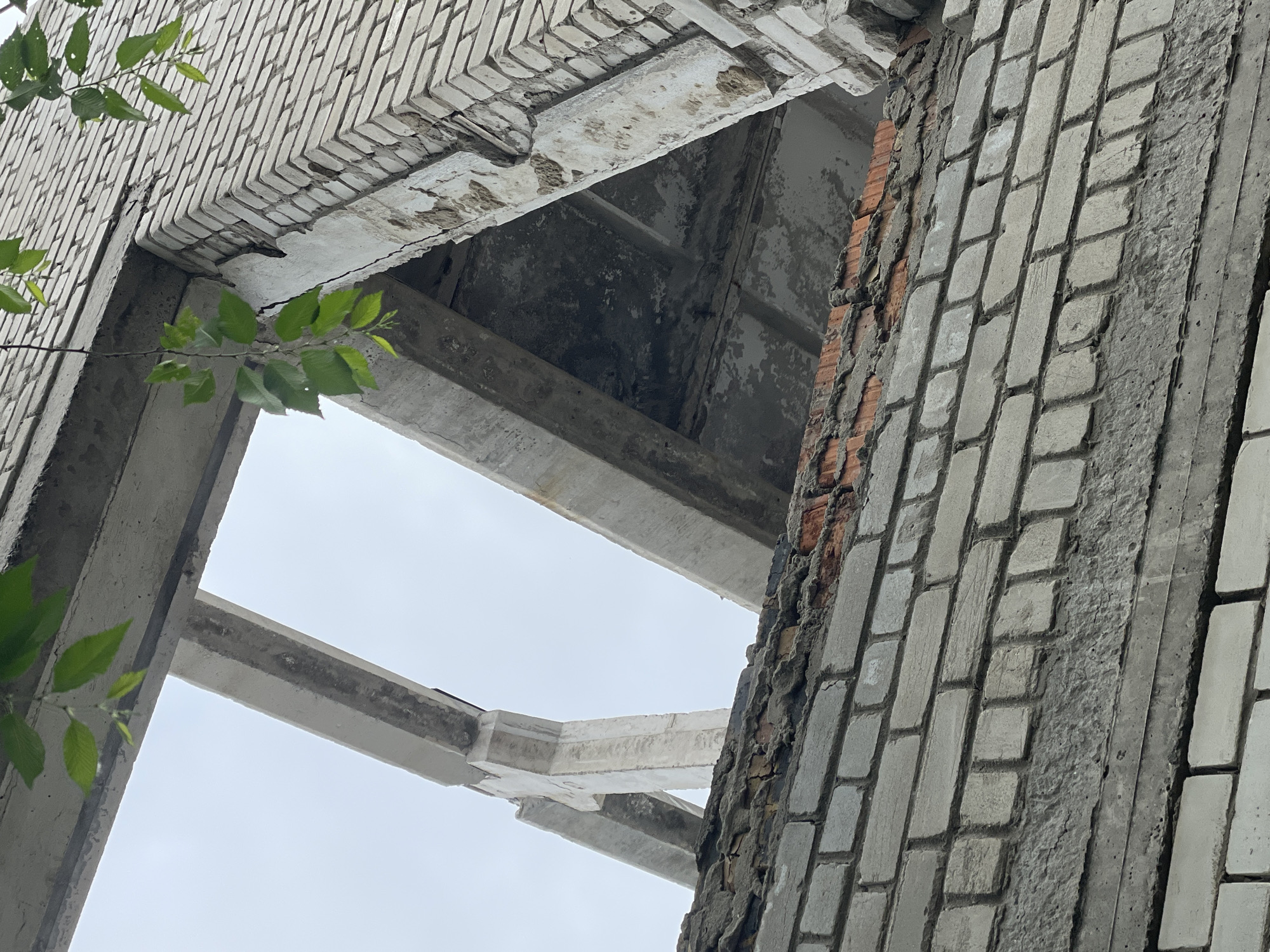 The image captures an upward view into the decaying structure of what appears to be an abandoned or derelict building. The scene is dominated by stark architectural elements made of concrete and brick, partially obscured by green foliage.
Concrete beams form a complex, angular framework, extending across the frame and creating openings through which a light grey, overcast sky is visible. These beams show significant signs of wear and deterioration, with rough textures, visible cracks, and sections where material has chipped away.
The walls are constructed from light-colored bricks, possibly a facade or rendered surface, laid in a distinctive pattern. On the right side of the frame, a vertical section of the wall reveals patches of darker, reddish brick underneath the lighter surface, suggesting damage or incomplete construction. The texture of the brickwork is uneven, with some bricks appearing eroded or covered in grime.
Thin branches with vibrant green leaves from a plant or tree intrude into the frame from the left and lower-left, providing a natural contrast to the industrial decay. These leaves are a notable detail, highlighting nature's reclamation of the structure.
No specific activity or event is taking place; the image portrays a static moment of architectural decay. The overcast sky suggests a cloudy day, contributing to the somewhat somber and desolate atmosphere. There is no visible text in the image.