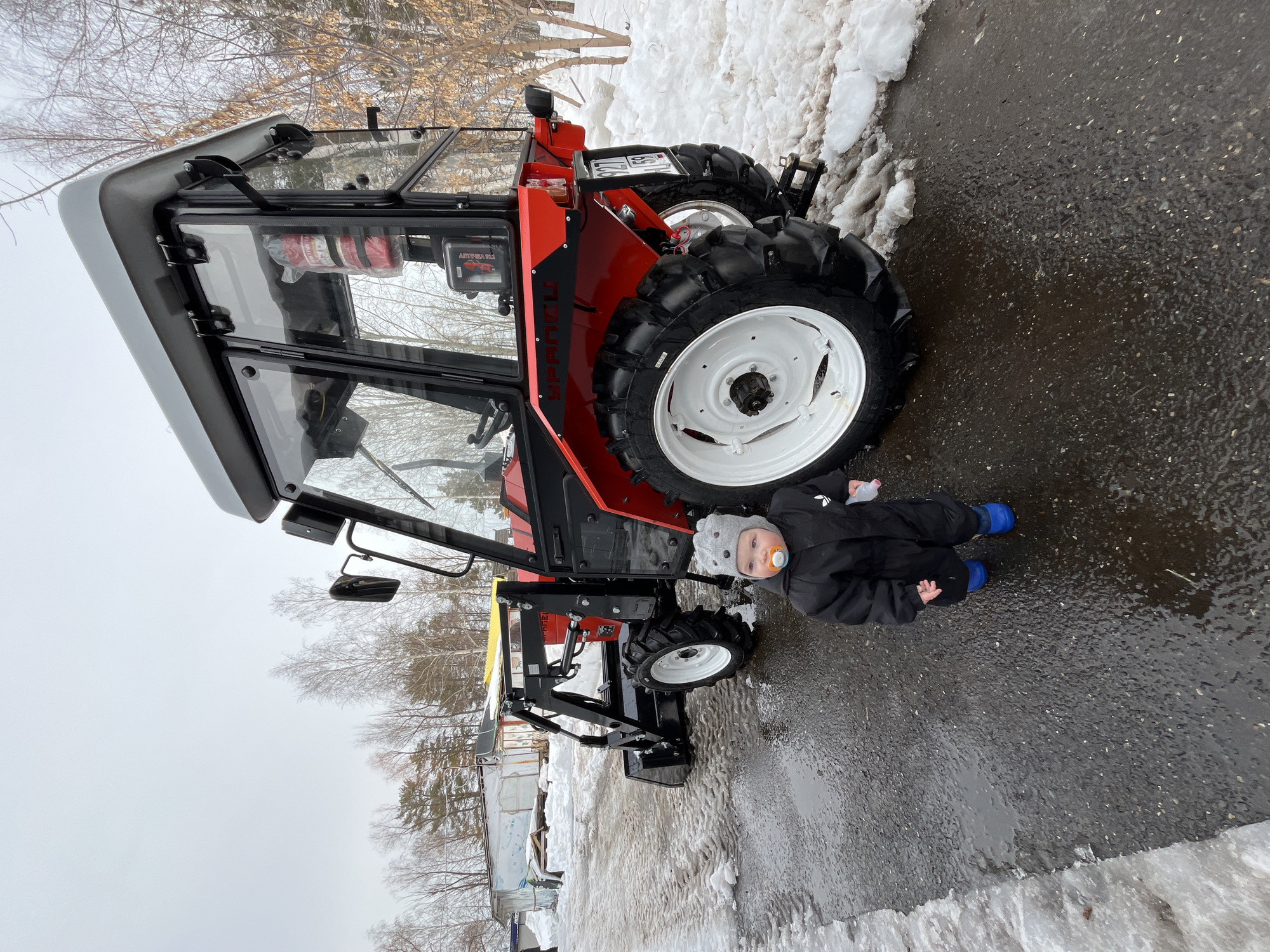 The image displays an outdoor scene featuring a small child and a compact utility tractor on a wet asphalt surface with melting snow. The setting is in Chaikovsky, Perm Krai, Russia, identified by the "59" regional code on the tractor's license plate.
A child, appearing to be a toddler, stands in the foreground, slightly left of center, wearing a grey knitted beanie, a black full-body snowsuit (with a small white logo, possibly Adidas), and blue boots. A pacifier is present in the child's mouth, and a clear plastic bottle with a pink cap is held in the right hand. The child faces forward, looking towards the viewer.
To the child's right, a red and black compact utility tractor is parked. It features a red main body, a black chassis, and a grey-roofed cabin with transparent windows. A red fire extinguisher is visible inside the cabin. The tractor is equipped with a black front-end loader and bucket attachment. It has large, knobby tires with white rims. A Russian license plate, "927 T1 | 59," is affixed to the rear. Black text spelling "УРАЛЕЦ" (URALETS) is visible on the tractor's red side panel above the rear wheel.
The ground is dark, wet asphalt, with patches of melting snow and slush along its edges, particularly on the left and in the background. Tire tracks are visible in the snow. The background shows an area with bare deciduous trees and some coniferous trees under an overcast, light grey sky. To the far left, several utilitarian structures, including a white and blue trailer or shed, are partially visible. No other individuals are present in the scene.
