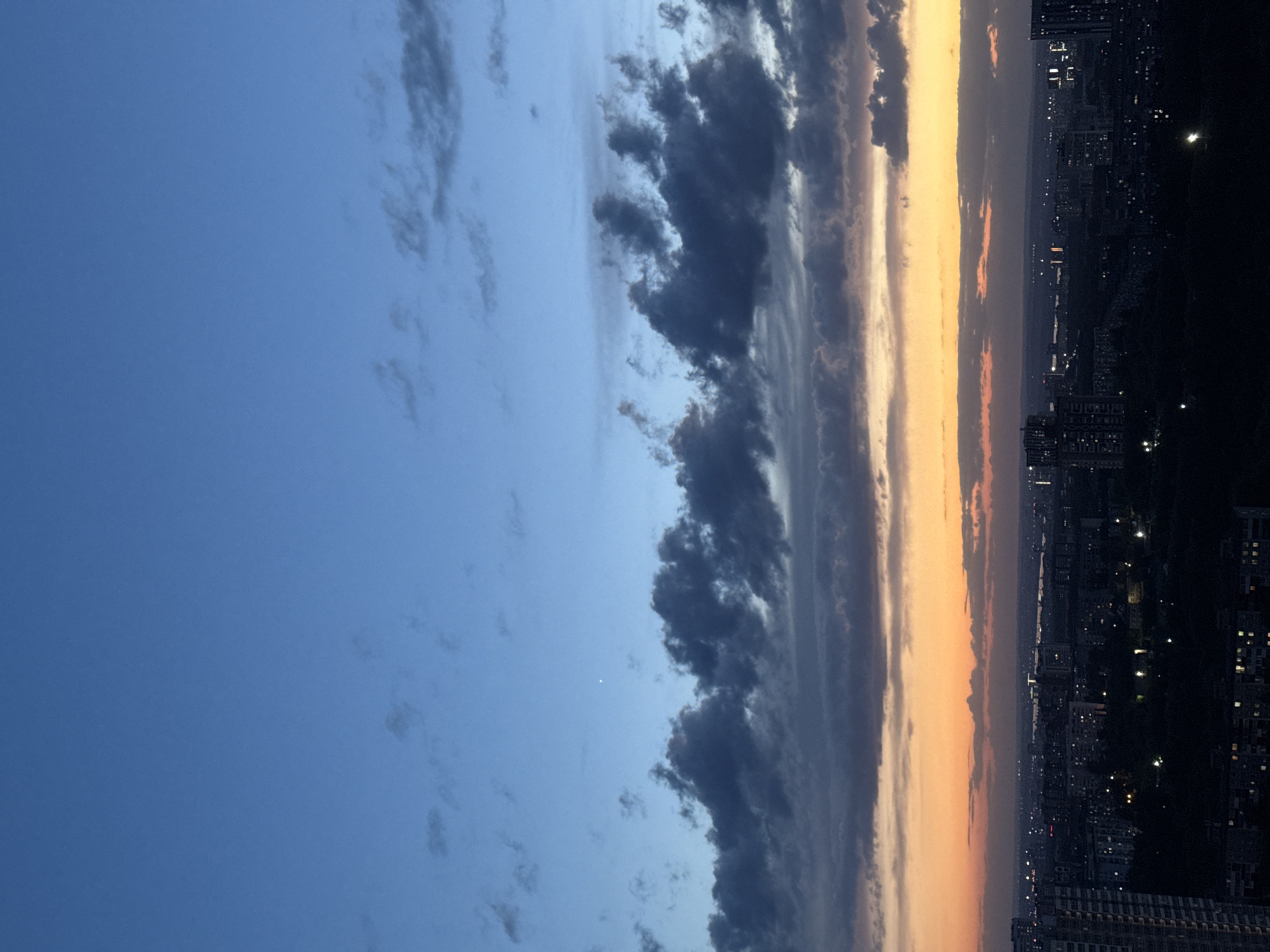The image depicts a twilight scene in Perm, Russia, showing a gradient sky transitioning from blue to orange, with a band of dark clouds separating the sky from a city skyline. The right side of the image shows a densely populated urban area with illuminated buildings against a dark background. The photograph is taken from an elevated position, potentially a high-rise building, capturing a wide view of the sky and the cityscape.