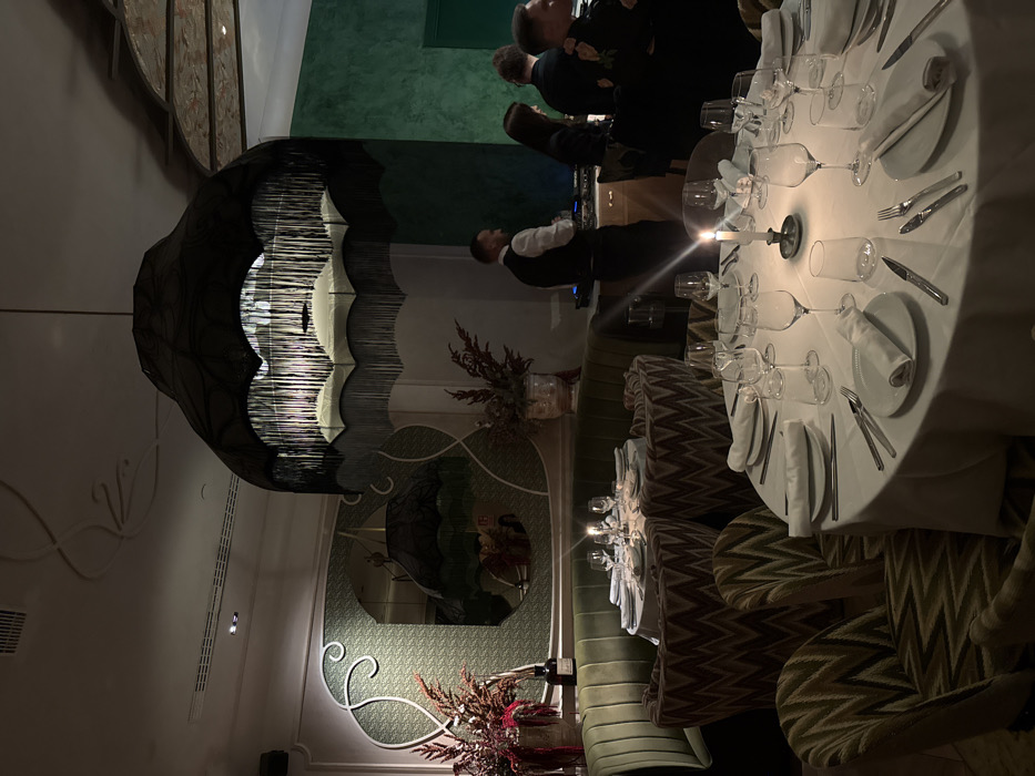 A dimly lit restaurant showcases a table elegantly set with white linens, multiple wine glasses, plates, and silverware. A lit candle centerpiece adds to the ambiance. The room features unique decor, including a large, stylized chandelier and a decorative mirror. Several individuals stand in the background. Zigzag-patterned chairs line the table, completing the upscale dining setting.