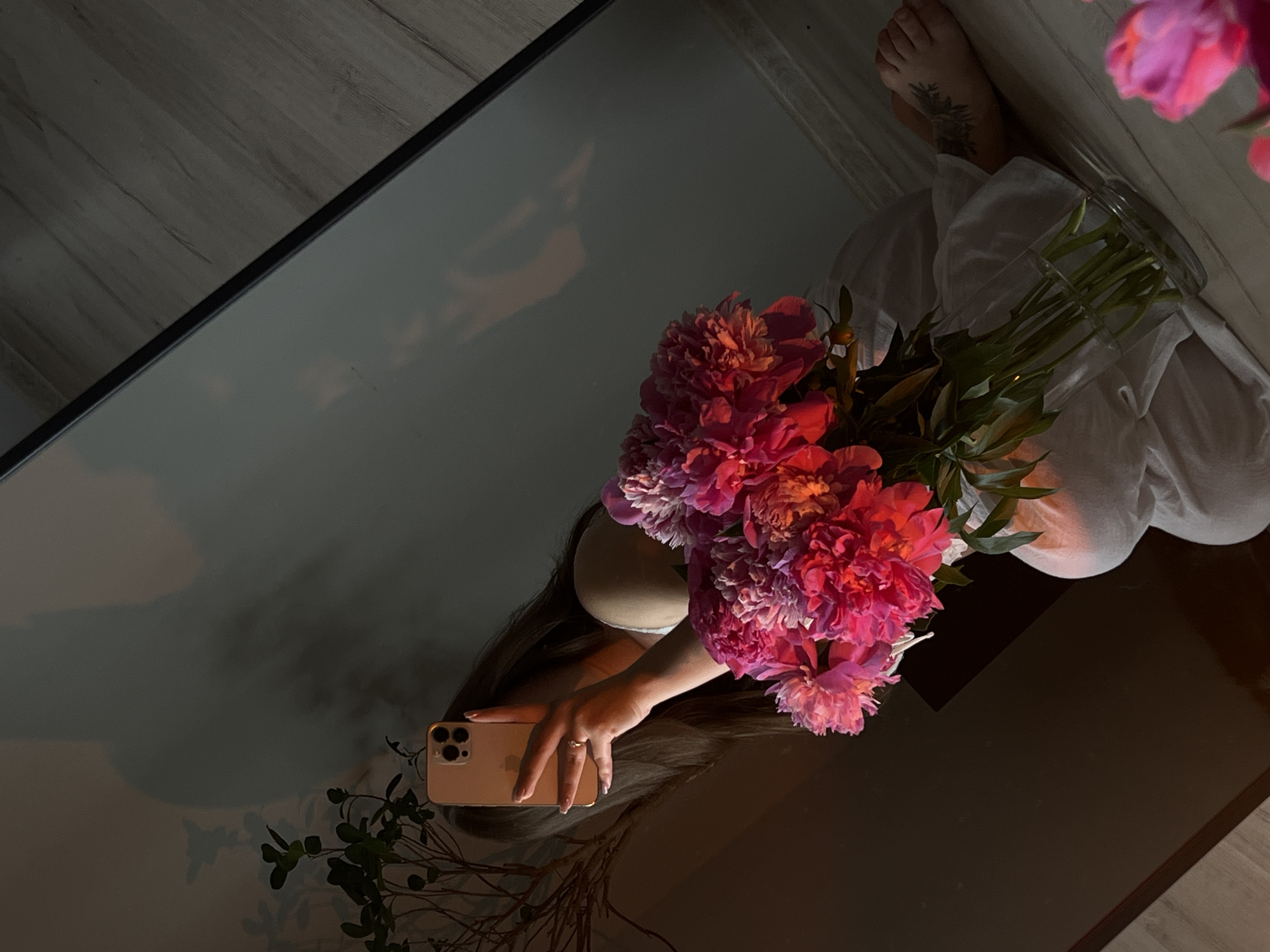 A person is taking a mirror selfie, holding a bouquet of pink peonies and a smartphone. The person is wearing a white dress and is seated on the floor. The background consists of a wooden floor and a wall. A plant is visible to the left. The image is taken indoors, and the lighting suggests it is either early morning or late evening. There is no indication of the city of Ternopil, Ukraine.