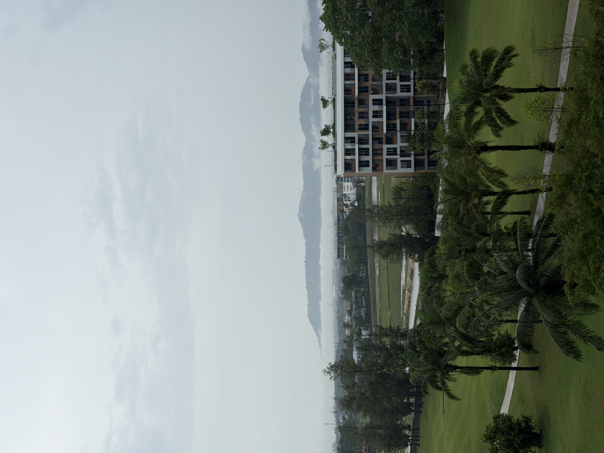 The image shows an overcast day in Choeng Thale, Thailand. A tall building with white and brown exterior walls is seen in the mid-ground. Palm trees are arranged along a path in a green grassy area, situated right of the building. A field can be seen further into the distance, past the building, and a mountain range runs along the horizon. The sky is filled with grey clouds.