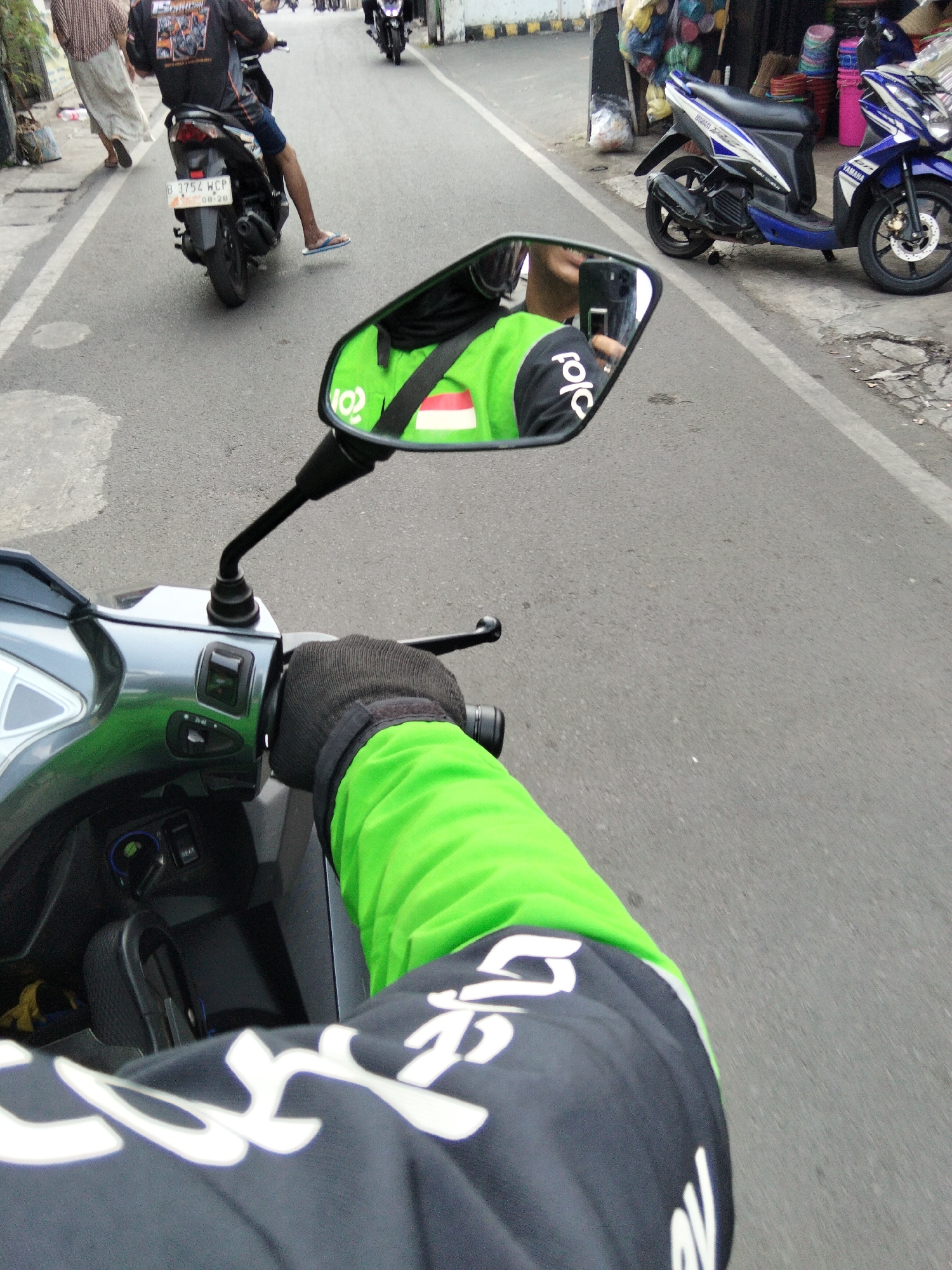 This is a first-person perspective image taken from a motorcycle in South Tangerang, Indonesia. The rider, wearing a black jacket with white lettering and a bright green long-sleeved shirt underneath, is operating the motorcycle. A gloved hand is visible on the handlebar. In the rearview mirror, the rider's face can be seen reflected, holding a smartphone.
Other motorcycles are present on the road, including one with a license plate that reads "B 3754 WCP". In the background, there is a roadside stall selling various items. A blue and white Yamaha motorcycle is parked on the sidewalk. The road appears to be paved and is in moderate condition with some cracks. The environment suggests a typical urban street scene in Indonesia.