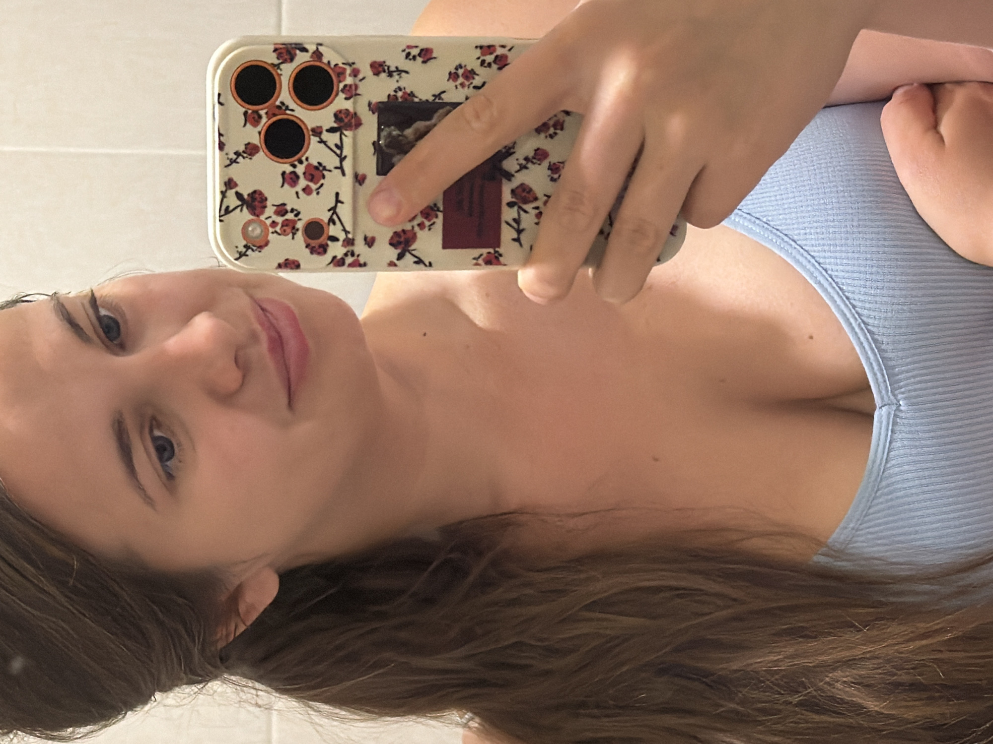 A young woman with long brown hair tied back in a ponytail is taking a selfie in a bathroom with white tiled walls. She is wearing a light blue ribbed tank top. She is holding a smartphone in her right hand, which has a floral case with red and white flowers. Her left hand is resting on the phone. The woman is looking at the camera with a slight smile. The lighting suggests it is indoors, possibly a domestic bathroom. The image does not contain any information that would place the location in Sochi, Russia.