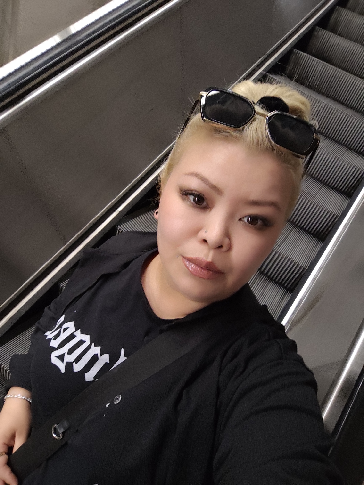A woman with short blonde hair pulled back in a bun, is standing on an escalator in an indoor setting. She is wearing black sunglasses on top of her head, a black t-shirt with white gothic-style lettering and a black overshirt. A black bag strap is diagonally across her chest. She has a nose piercing and is wearing makeup, including eyelash extensions. The escalator is made of grey metal with black ridges. A metallic railing runs alongside the escalator. The location is not specified to be in Sarıyer, Turkey, so that is not included in the description.
