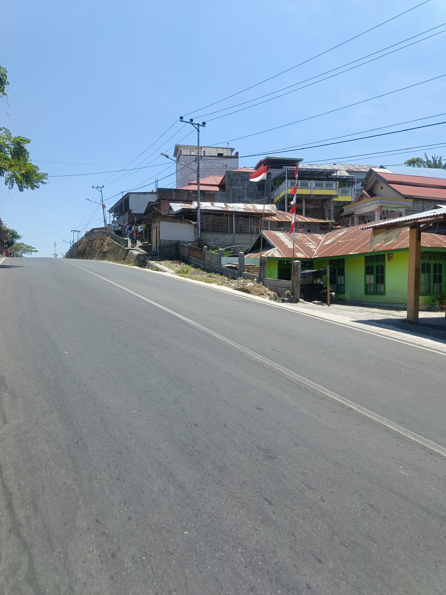 A street scene in Buhu, Indonesia, captures a bright, sunny day with a clear blue sky. A paved road ascends a gentle incline, with a white line marking its center. On the right side of the road, a row of buildings lines the street. The closest building is painted a vibrant lime green with several windows featuring green frames. It has a rusted corrugated metal roof and a wooden pillar supporting an overhang. Behind this building, other structures are visible, including some with red tiled roofs and others with corrugated metal roofs. Several Indonesian flags, red and white, are planted in front of some of the buildings.

Further up the hill, more buildings are clustered together. These appear to be a mix of residential and possibly commercial properties, with varying architectural styles and materials. Some buildings have multiple stories. Utility poles with electrical wires are present along the street and extending over the buildings. A few people are visible walking on a raised area beside the road, near some of the buildings. A small, dark-colored vehicle, possibly a motorcycle with a sidecar, is parked in front of the green building. The vegetation on the left side of the road consists of trees and some dry grass on the embankment. The overall impression is of a roadside community on a warm day.