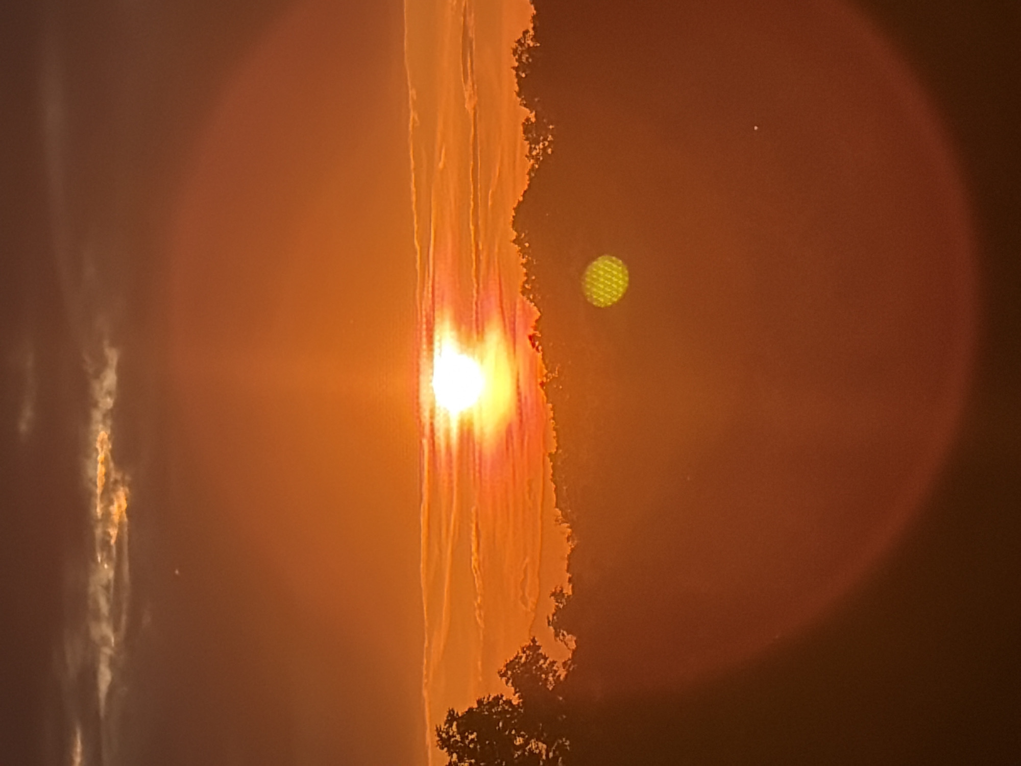 The image shows a sunset, obscured by what appears to be smoke and atmospheric distortion. A bright sun is prominently positioned slightly off-center. An orange glow envelops the scene. An area of land covered in trees and bushes is barely visible at the bottom of the frame. A patterned, yellowish-green object is also visible. A bright pinpoint of light is on the opposite side of the orange glow. The photograph does not provide enough information to determine the location within Plovdiv. There are no indications of people or specific actions. The scene seems to be outdoors, focused on the sunset.