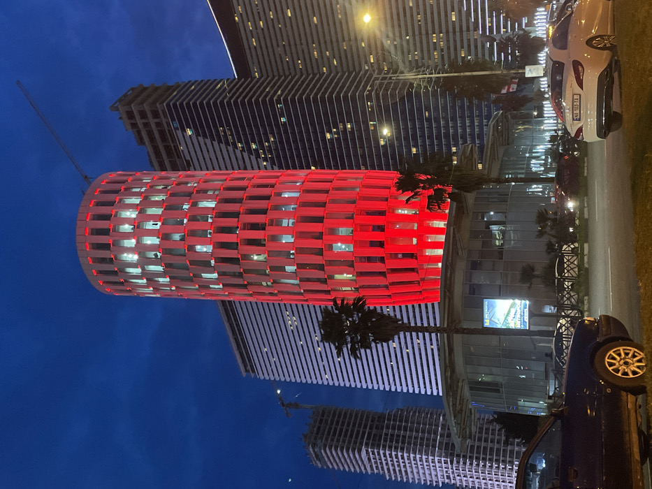 Buildings dominate the image, one a cylindrical structure illuminated in red. Adjacent are taller, linearly designed buildings reflecting streetlights. Cars line the right edge, partially obscuring the view. Palm trees punctuate the scene, adding vertical elements. The sky transitions from dark blue to black. A billboard and construction crane are subtly visible in the background.