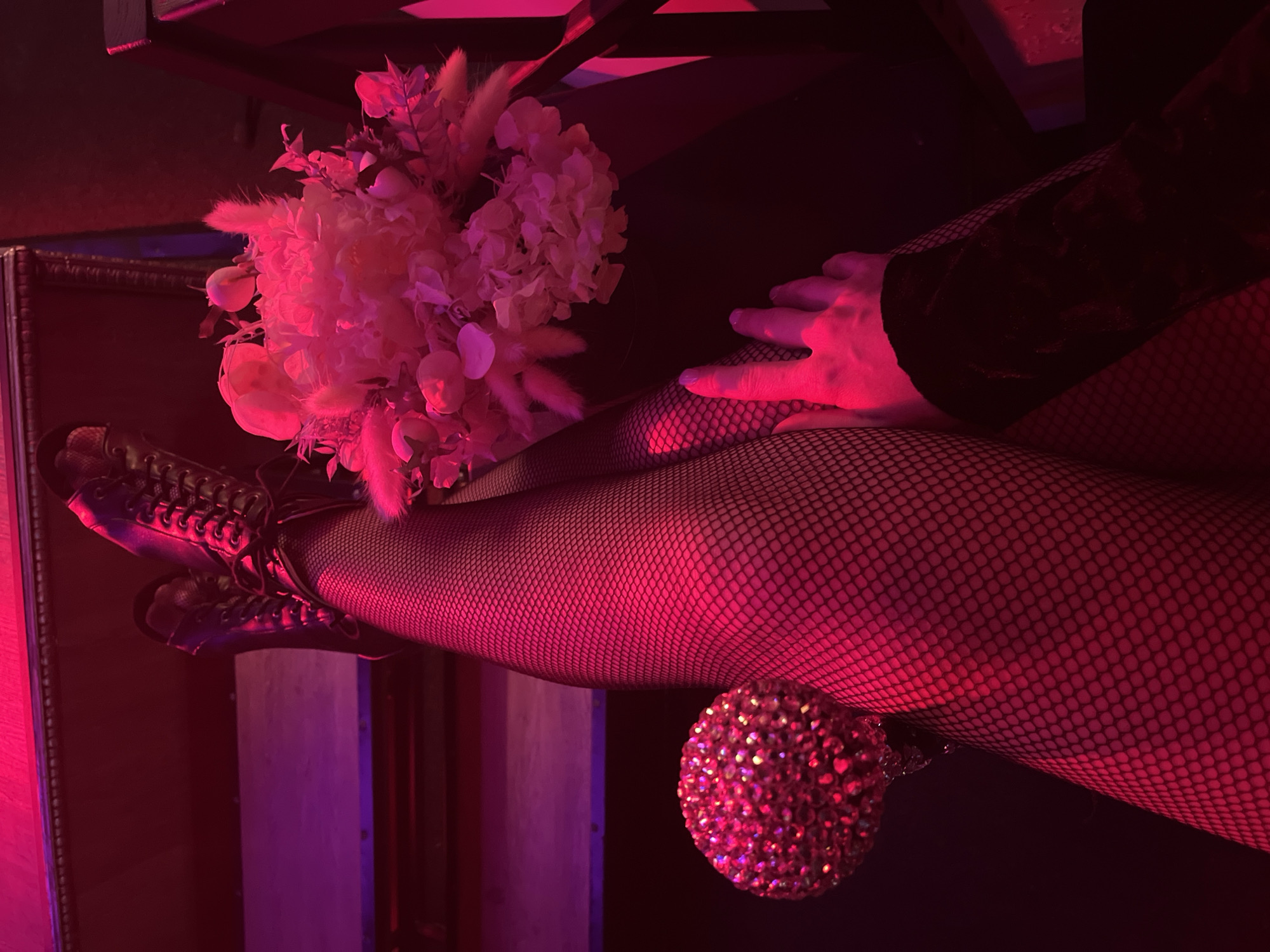 In an indoor setting, likely a dimly lit room with red and purple lighting, a person's legs and feet are prominently displayed. They are wearing black fishnet stockings and black lace-up boots. One leg is bent at the knee, with the foot resting on a lower surface. The other leg is extended. To the right of the legs, a bouquet of dried flowers, predominantly pink and beige, is arranged in a dark container. To the left of the legs, a round, red, sequined object is visible. A hand with painted nails is resting on the fishnet stocking of the extended leg, with fingers pointing downwards. The background includes dark surfaces and what appears to be a tiered structure, possibly stairs or shelves, illuminated by the colored lighting. The overall atmosphere is moody and theatrical. The image does not contain any elements that would allow for the identification of the city as Kazan, Russia, or any specific location within it.