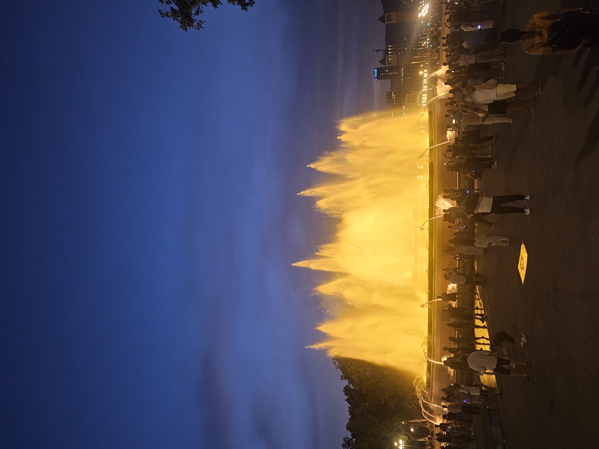 The image shows the Magic Fountain of Montjuïc in Barcelona, Spain, at dusk. A large, illuminated fountain is the central focus, with water jets shooting upwards and glowing a bright yellow. Numerous people are gathered in front of the fountain, standing on a paved area. Many are holding up phones to take pictures or videos of the spectacle. In the background, beyond the fountain and trees, are buildings with lights on, and a tall tower. The sky is a deep blue with some faint clouds. The overall scene is one of public enjoyment of a light and water show.