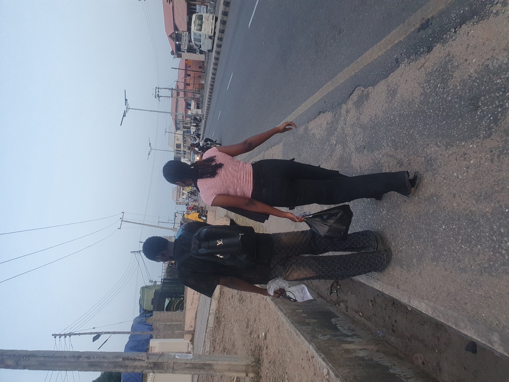 Two individuals are walking along the roadside in an urban area of Lokoja, Nigeria. One person, possibly female, is wearing a light pink striped shirt and dark trousers. The second person, possibly male, is wearing a black shirt, patterned trousers, and carrying a black backpack. Both are holding plastic bags. They are walking alongside a road with moderate vehicle traffic. Buildings and utility poles are visible in the background. The sky is overcast.