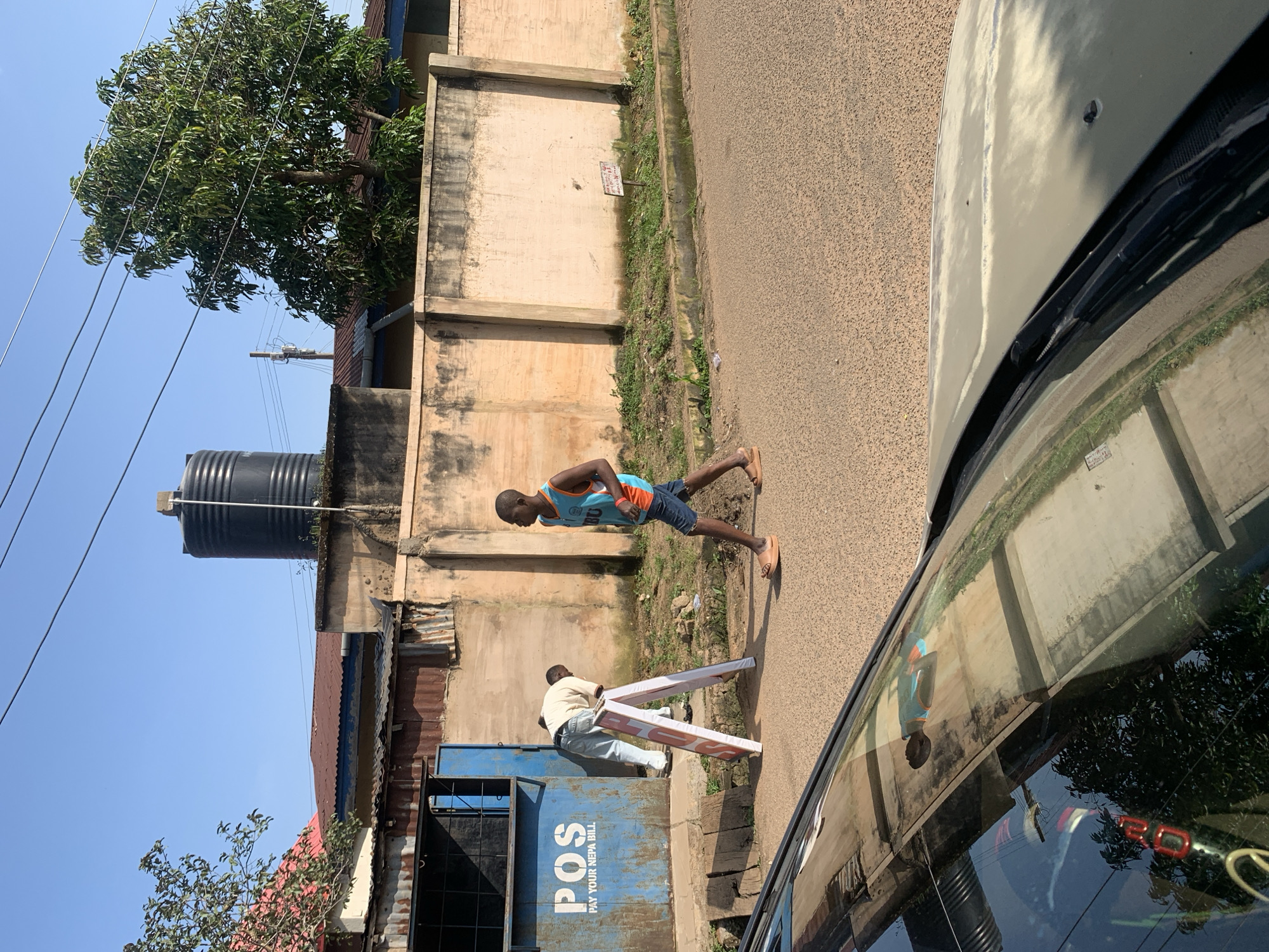 A young Black male wearing a blue tank top and denim shorts walks on a paved road in Jos, Nigeria. He is walking to the right of the frame. He has on sandals. Behind him, a Black male wearing a light yellow shirt and light blue pants bends over a white A-frame sign. The sign has red writing on it. To the left of the sign is a blue structure with the white letters "POS" and "PAY YOUR NEPA BILL" on it. The structure appears to be a shop. Behind the shop, there are buildings with corrugated metal roofs. A large black water tank is visible on top of one of the buildings. A large tree with green leaves is to the right of the frame, behind a pale yellow wall. The sky is clear and blue. The foreground of the image is dominated by the reflection on a car windshield, showing the road, buildings, and trees.