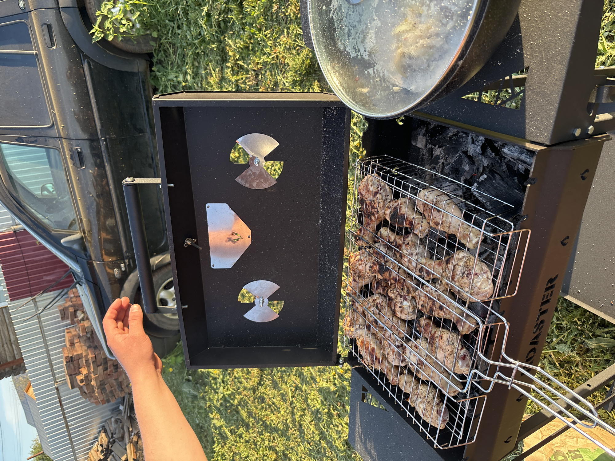 The image shows a barbecue grill in an outdoor setting, seemingly in a grassy area. The grill contains a wire basket filled with what appears to be chicken pieces, partially cooked and slightly charred. Underneath the chicken, there are visible charcoal pieces. The grill has a lid, partially visible, and some adjustable vents. To the left of the grill, a black car is seen, suggesting the presence of a residential area. There is a hand partially visible in the lower left corner of the frame. The image is rotated counterclockwise. The location appears to be Фоки, Russia, based on the metadata. The environment is natural with grass and the presence of a car and possible building supplies (bricks) nearby.