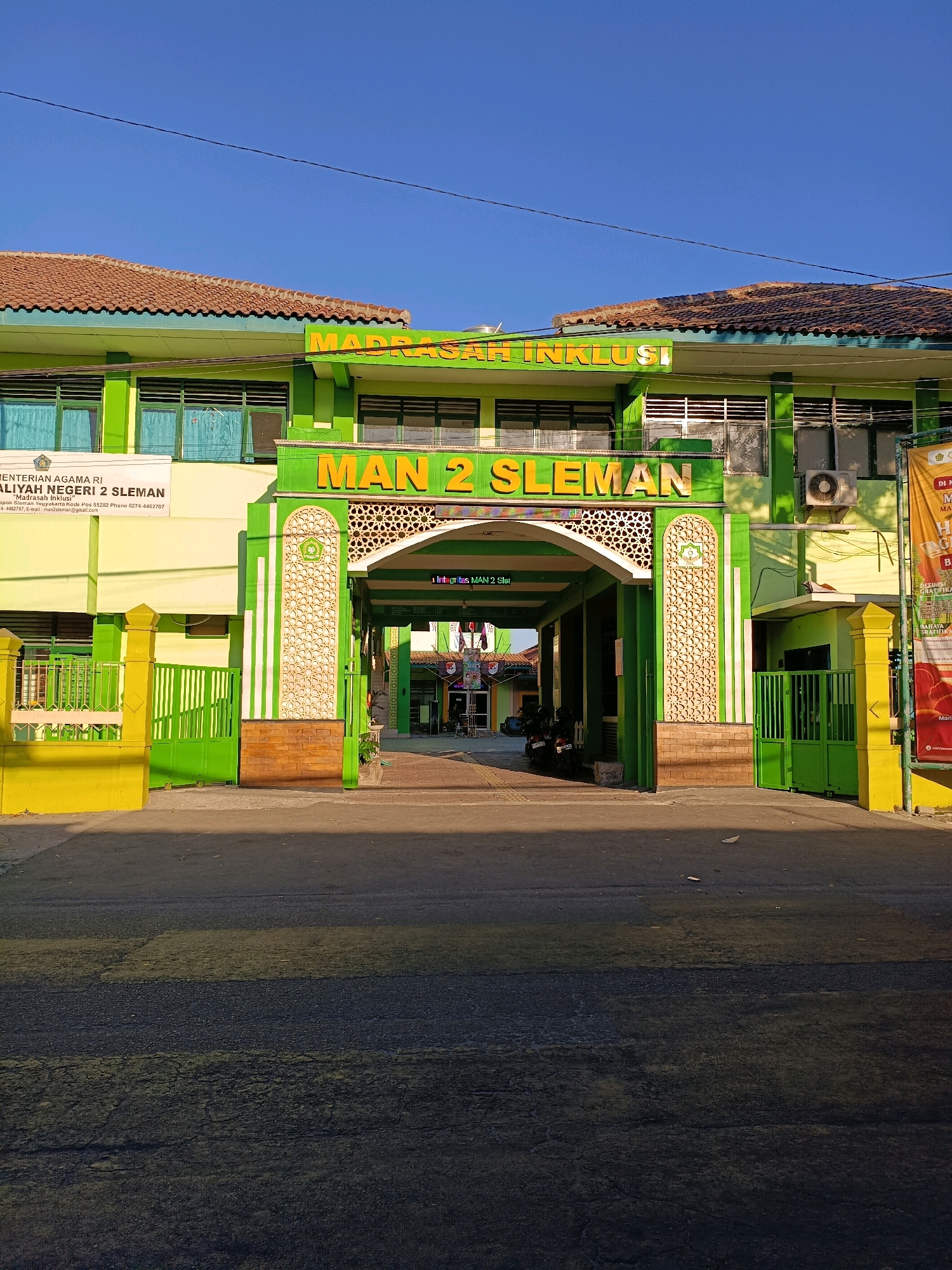 The image shows the entrance to Madrasah Aliyah Negeri (MAN) 2 Sleman, a public Islamic high school located in Maguwoharjo, Indonesia. The school building is painted in bright green with orange lettering that reads "MADRASAH INKLUSI" and "MAN 2 SLEMAN". The entrance is an archway decorated with Islamic geometric patterns and flanked by green gates. A banner with the school's name and logo is visible on the left side of the entrance. The sky is clear and blue, indicating a sunny day. The foreground shows a paved road with a pedestrian crossing. There are a few motorcycles parked inside the school grounds, suggesting it is a normal school day. The overall scene depicts a well-maintained educational institution in a tropical climate.