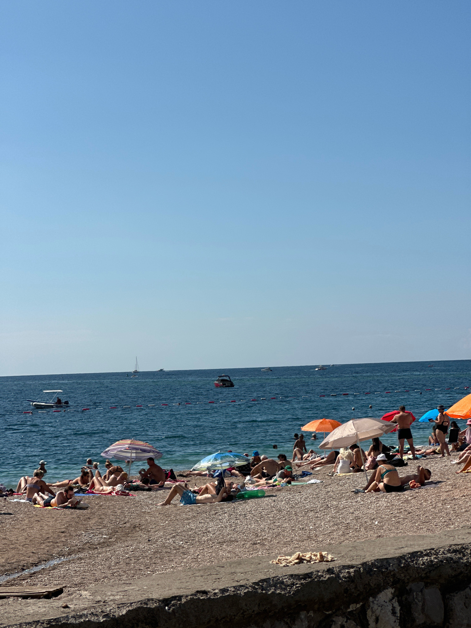 The image depicts a crowded beach scene on a sunny day, likely in Budva, Montenegro, given the context. The foreground is a pebble beach with numerous individuals sunbathing and relaxing on towels and blankets. Several colorful umbrellas provide shade. In the middle ground, the blue sea is dotted with several boats, including a small motorboat with a person at the helm, sailboats, and other recreational vessels. A rope barrier with buoys separates the swimming area from the open water. The background is dominated by a clear, light blue sky. The overall atmosphere is one of leisure and summer vacation.