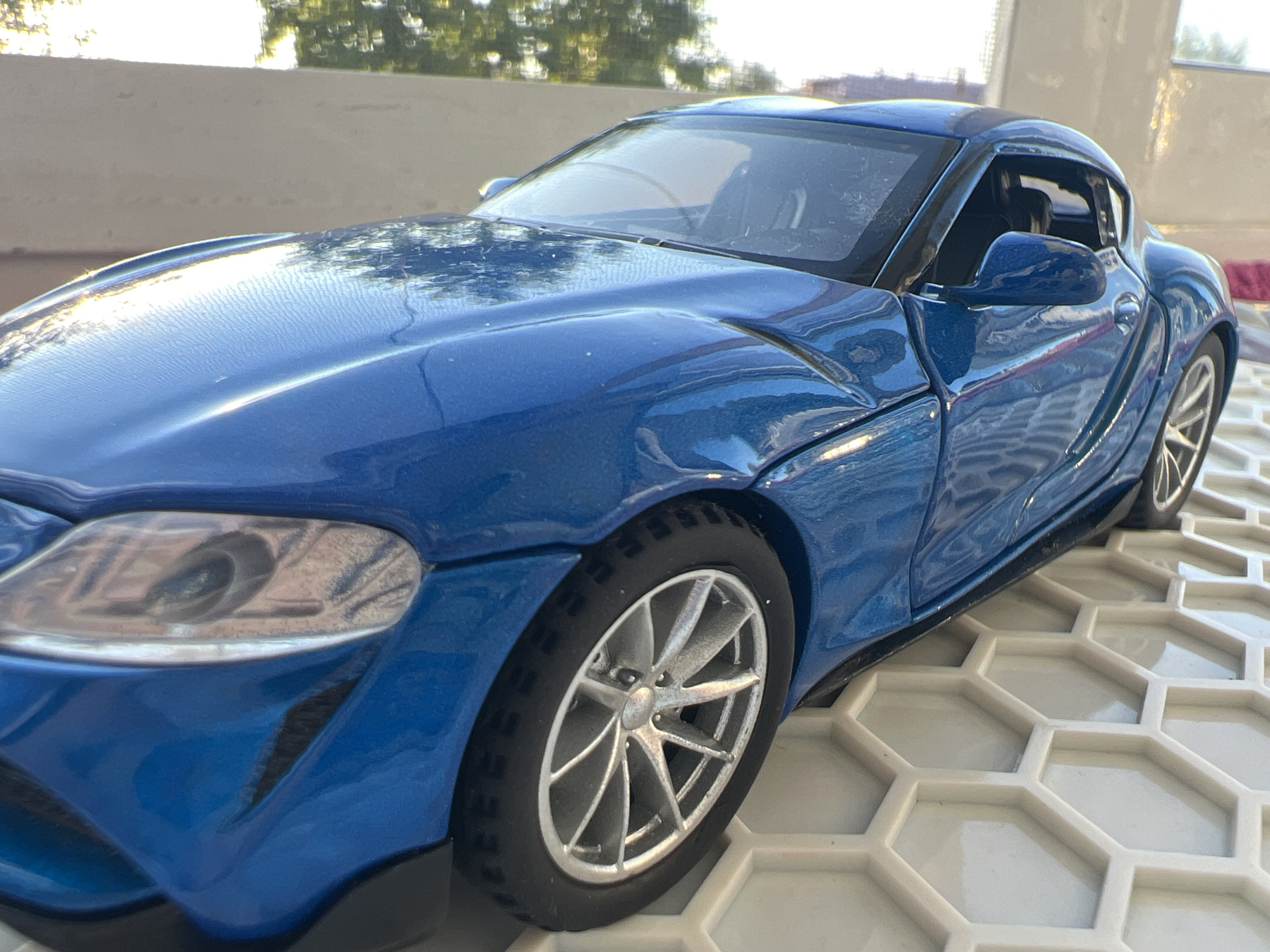 A blue toy sports car is on a hexagonal white surface, with a textured wall and blurred trees in the background. The car is positioned diagonally, facing towards the left side of the image. The front of the car, including the headlight and hood, is visible, as is part of the passenger side door and rear wheel. The scene appears to be outdoors, possibly on a balcony or patio, under natural lighting. There are no people or animals present in the image, and no specific details suggest a location in Omsk, Russia.