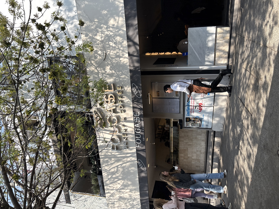 A daytime outdoor shot captures the front of a bakery and coffee shop, "FOUGASSE." A tree with sparse leaves is on the left. The building's white facade contrasts with the dark, receding entrance, where a worker in an apron stands. A group of people is on the sidewalk in front of the entrance. An advertisement for internet service is visible on the right of the doorway. The image is slightly rotated.