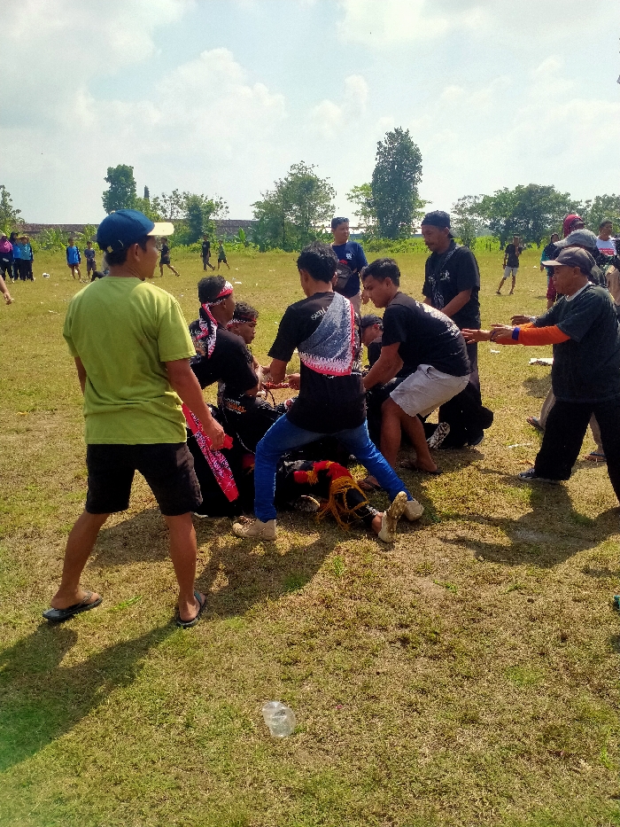 The image depicts an outdoor scene on a dry grass field in Celep, Indonesia, under a bright, partly cloudy sky, with strong sunlight casting distinct shadows. In the center-right, approximately ten adult males are actively engaged with a long, dark, cylindrical object, partially resting on the ground. This object features red and gold/yellow fringed or tasseled decorations on its visible end. Participants, dressed in a mix of casual attire including black t-shirts (one with "Seni" and a graphic print on the back), blue jeans, and shorts, are physically pulling and pushing the object. One participant wears a red and white patterned headband. To the left foreground, a male in a light green short-sleeved shirt, dark shorts, a blue baseball cap, and flip-flops stands observing the central activity, holding a red and white patterned cloth in his right hand. A crumpled, clear plastic bottle lies on the grass in the lower center foreground. The background shows numerous additional individuals scattered across the expansive field, with trees and distant structures visible on the horizon.