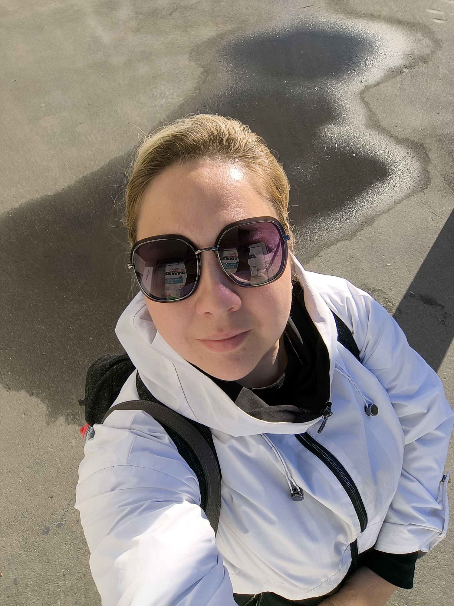 An overhead, close-up, selfie-style shot shows a woman with blonde hair pulled back, wearing large, dark-rimmed sunglasses and a white hooded jacket. The woman's face is turned slightly to her left, and her lips are slightly parted. A reflection of a white carton with green text is visible in the right lens of her sunglasses. The woman is wearing a black t-shirt underneath the white jacket, and a dark backpack strap is visible over her left shoulder. The background consists of a grey asphalt surface with irregular dark patches, possibly water or oil stains, and a distinct shadow cast from the right, suggesting an outdoor environment with sunlight. The scene is unidentifiable as a specific city location.