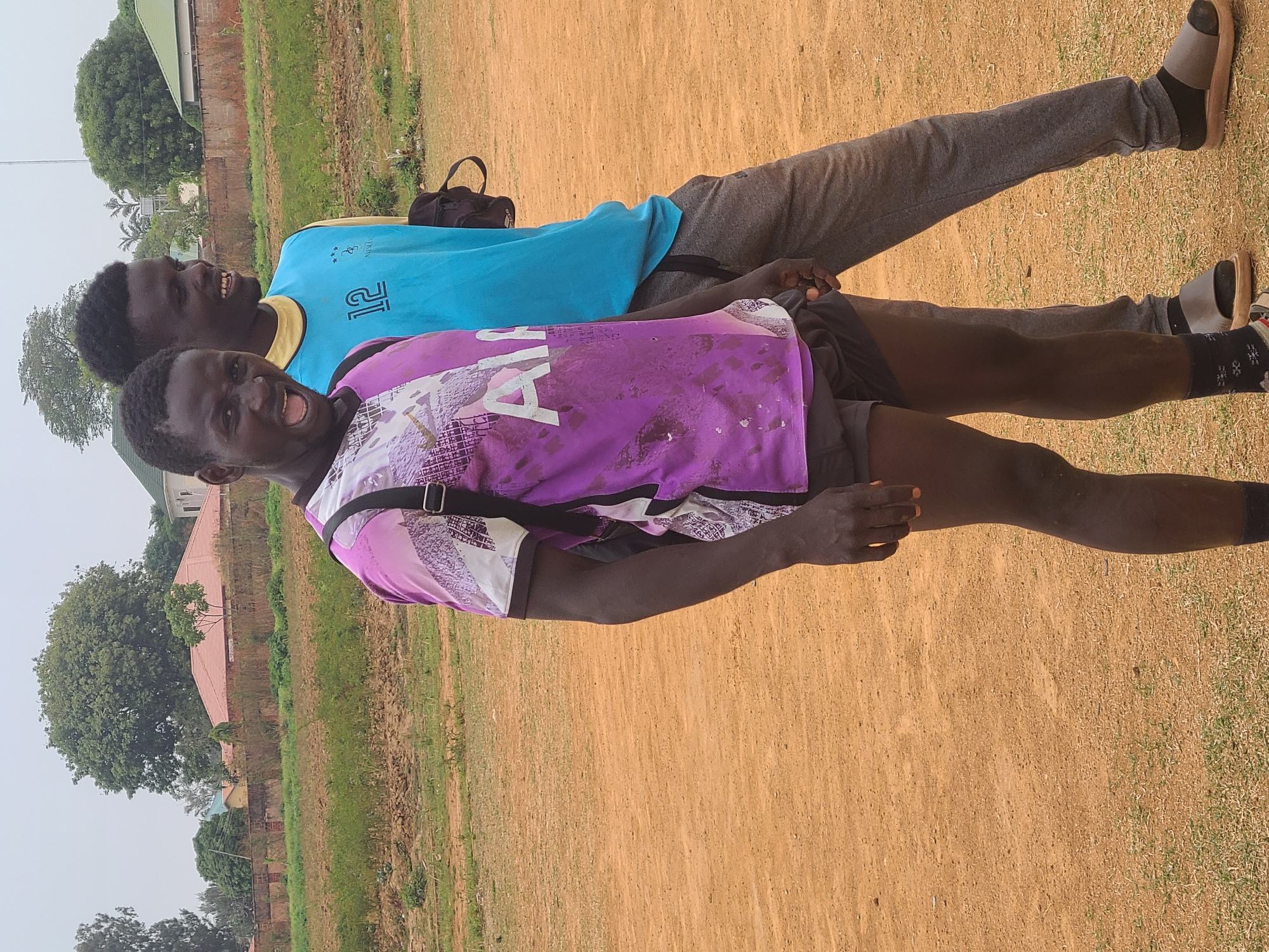 Two men are photographed outdoors in Barikin Jirga Summa, Nigeria. The foreground is a dirt field with some scattered green grass. In the background, there are buildings and trees. One man is wearing a purple and white patterned short-sleeved shirt, dark shorts, dark socks with white patterns, and brown sandals. He has a strap across his chest and is smiling with his mouth open. The other man is wearing a light blue short-sleeved shirt with yellow trim and the number 12 on the front, gray pants, dark socks, and dark sandals. He is also smiling. They appear to be standing close together. A black bag is visible in the background between them.