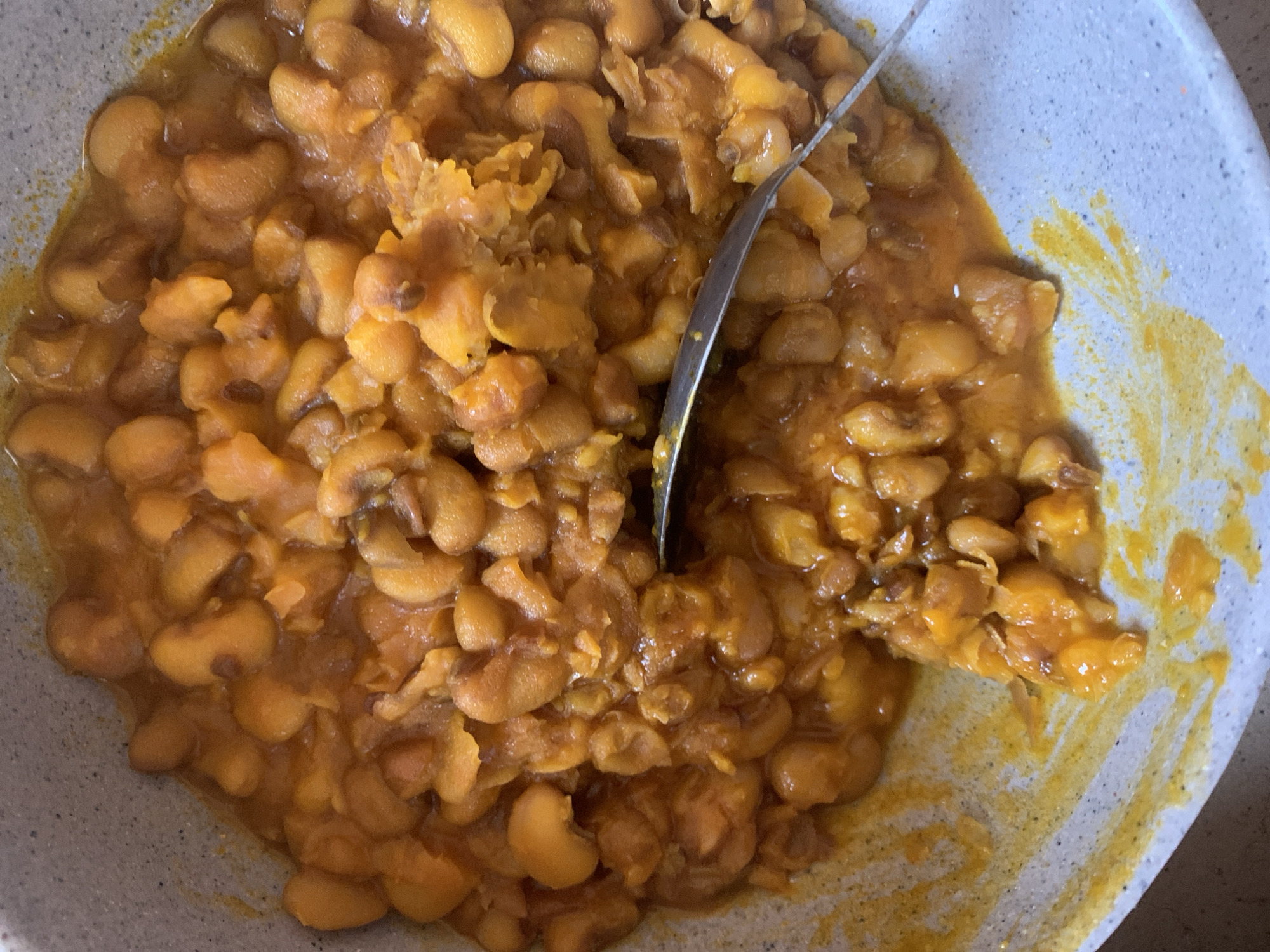 A top-down close-up shot shows a light grey speckled bowl filled with cooked brown sugar baked beans. The beans are a golden brown color and are coated in a thick, syrupy sauce that is also golden brown. Some of the beans are whole, while others are broken or mashed. A silver spoon is visible in the lower right corner of the bowl, partially submerged in the beans. The sauce has dripped over the side of the bowl onto the counter in a few places.