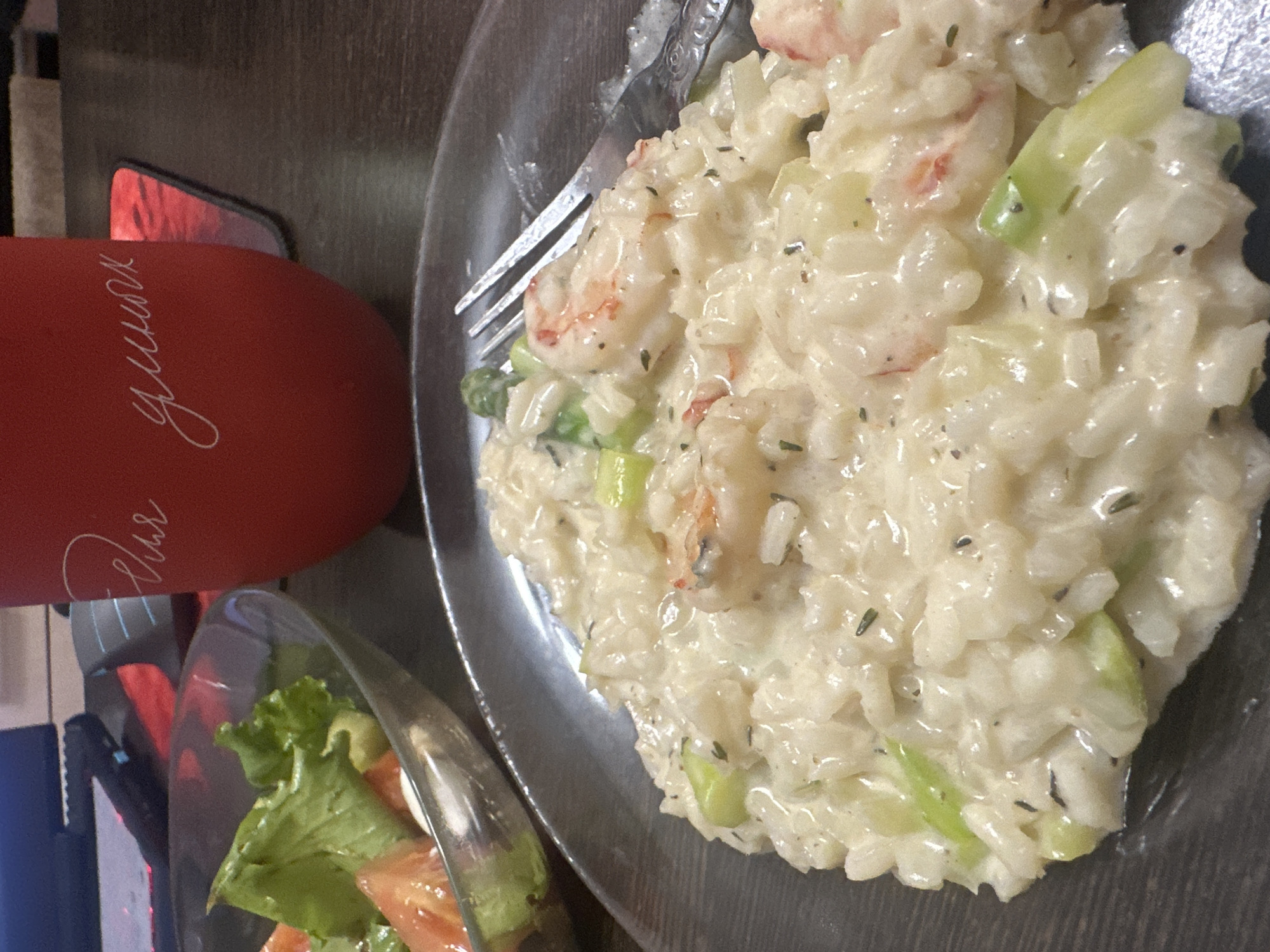 The image shows a close-up of a plate of shrimp risotto, served in a dark gray dish. The risotto is creamy and appears to be made with rice, shrimp, and green vegetables, possibly asparagus. A fork is visible, resting on the dish, with some of the risotto on its tines. To the left of the risotto, a small salad is visible in a clear glass bowl. The salad contains lettuce and tomato.
In the background, a red mug with white script lettering is partially visible. The text on the mug appears to be in Cyrillic script and reads "Для тебя". This translates to "For you". The overall scene suggests a meal is being prepared or enjoyed, likely indoors due to the lighting. The setting appears to be a home or a casual dining establishment. No specific time of day or weather conditions are discernible. The image is likely taken in Novoaltaysk, Russia, as indicated by the location context.