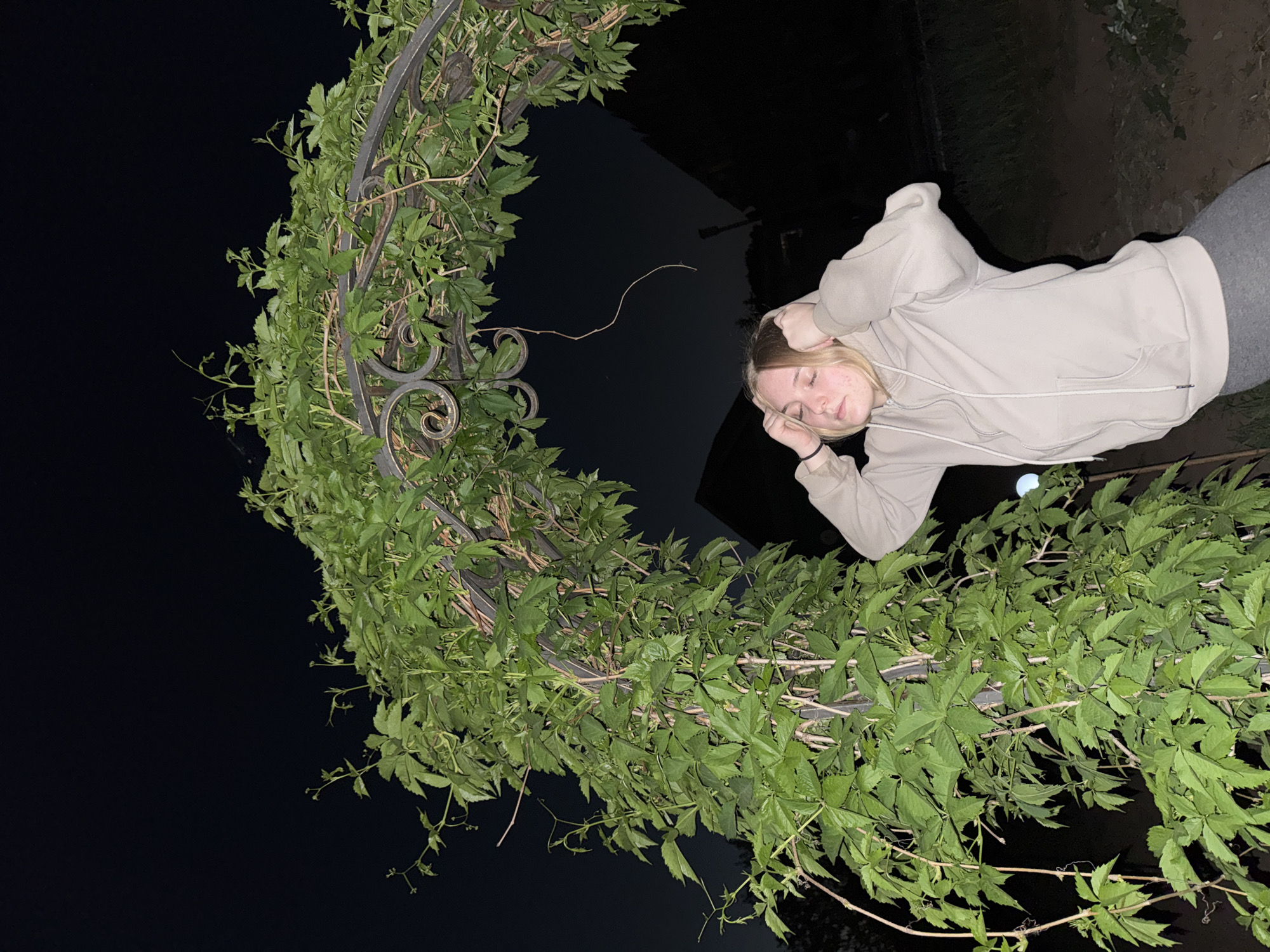 A woman with blonde hair is standing next to a metal arch covered in green plants. She is wearing a light beige hoodie and gray pants. Her eyes are closed, and she has her hands on her head. The background is dark, suggesting it is nighttime. The location is Zhigalki, Russia.
