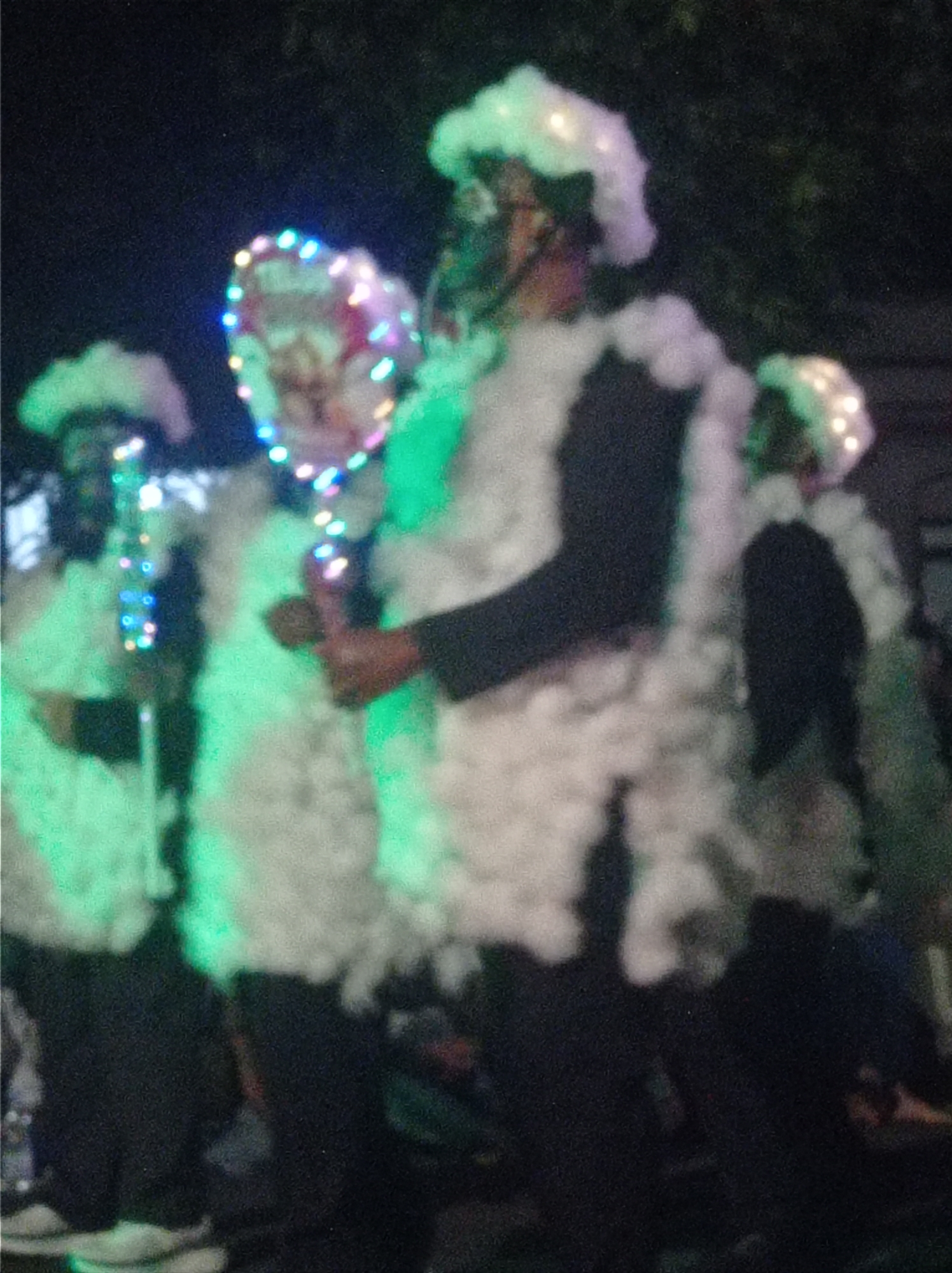 In Pandean, Indonesia, an undated image captures a nighttime scene featuring several individuals adorned in elaborate, fuzzy white costumes with festive lights. One person in the foreground, centrally positioned, wears a tall, plush white headpiece that partially obscures their face. They are also draped in a voluminous white, feather-like material that extends down their body and arms, resembling a large, fuzzy coat. This individual holds a heart-shaped object decorated with colorful, twinkling lights. To the left, another person in a similar white costume can be seen, also featuring the fuzzy white material and lights. Further to the left, a third individual in a white costume is visible, holding a vertical string of colorful lights. The overall impression is of a festive parade or performance, with the bright lights contrasting against the dark, nighttime environment. The background is dark and somewhat indistinct, suggesting an outdoor setting at night.