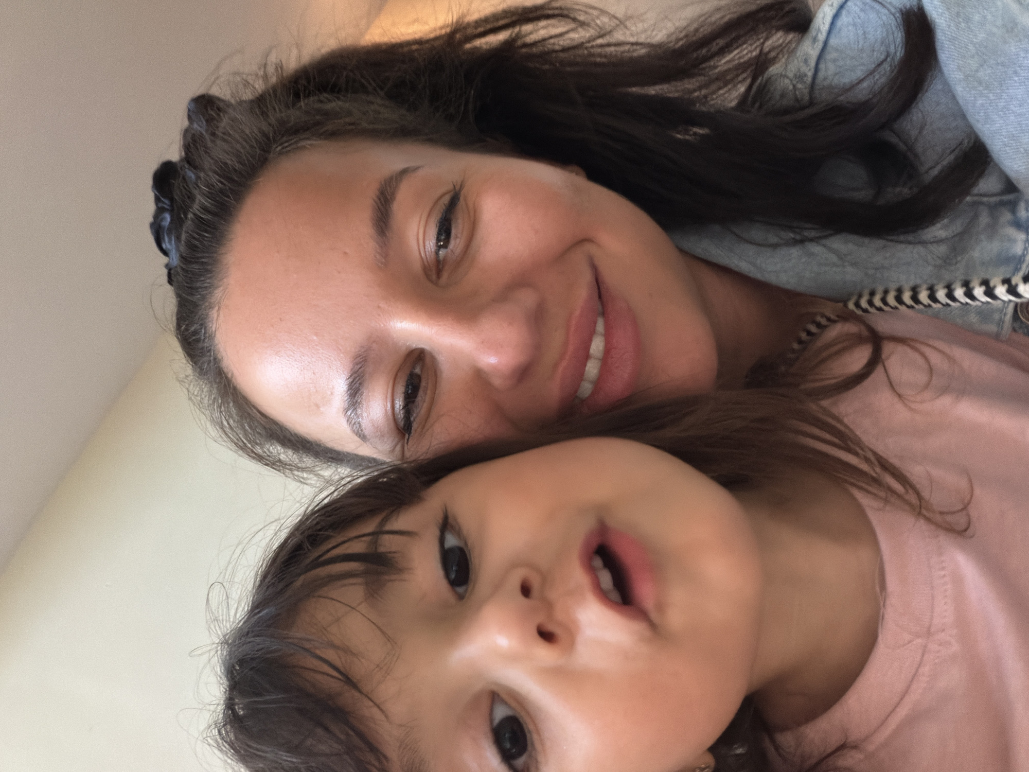 The image is a close-up selfie featuring two females, an adult and a child, positioned in the foreground. The adult, on the right, has dark hair tied up with a dark scrunchie, brown eyes, and is smiling with her mouth open, revealing teeth. She is wearing a light blue denim jacket. The child, on the left, has dark hair falling across her face, dark eyes, and her mouth is slightly open, appearing to be speaking or making a facial expression. She is wearing a pink t-shirt. The background is a plain, light-colored wall. The photo appears to have been taken indoors. There is no visible indication of the city of Burgas, Bulgaria, in the image.