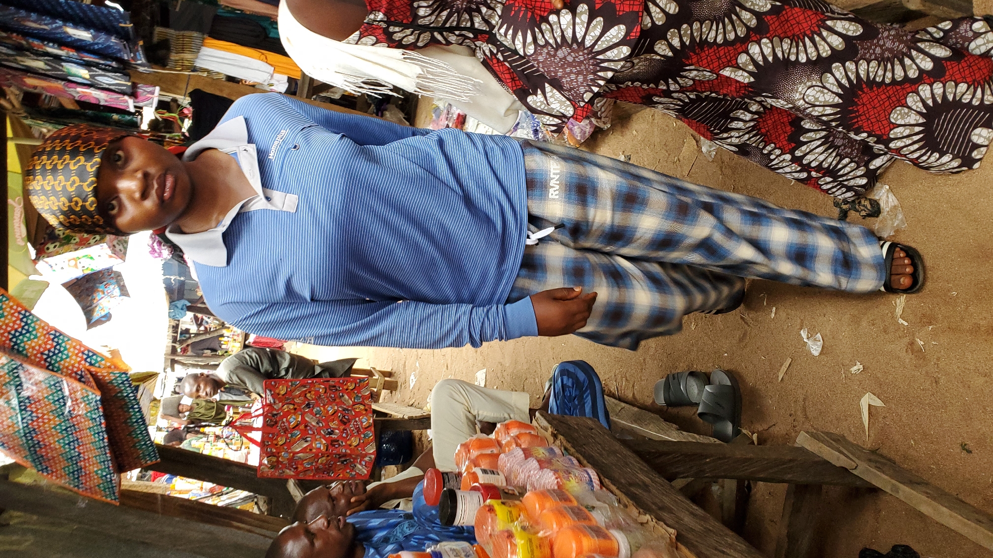 A medium shot captures a crowded outdoor market scene in Jalingo, Nigeria. The foreground is dominated by a young woman with dark skin and a patterned headscarf, wearing a blue striped long-sleeved shirt and plaid pants. She is walking towards the viewer, her expression neutral. To her left, a wooden stall displays rows of orange plastic containers, possibly cosmetics or food products. Behind her, other individuals are visible, including two men sitting at the stall and a man standing further back in a green garment. The background is filled with market stalls adorned with colorful fabrics and clothing items hanging for sale. The ground is covered in reddish-brown dirt. The overall atmosphere suggests a busy, typical market day.